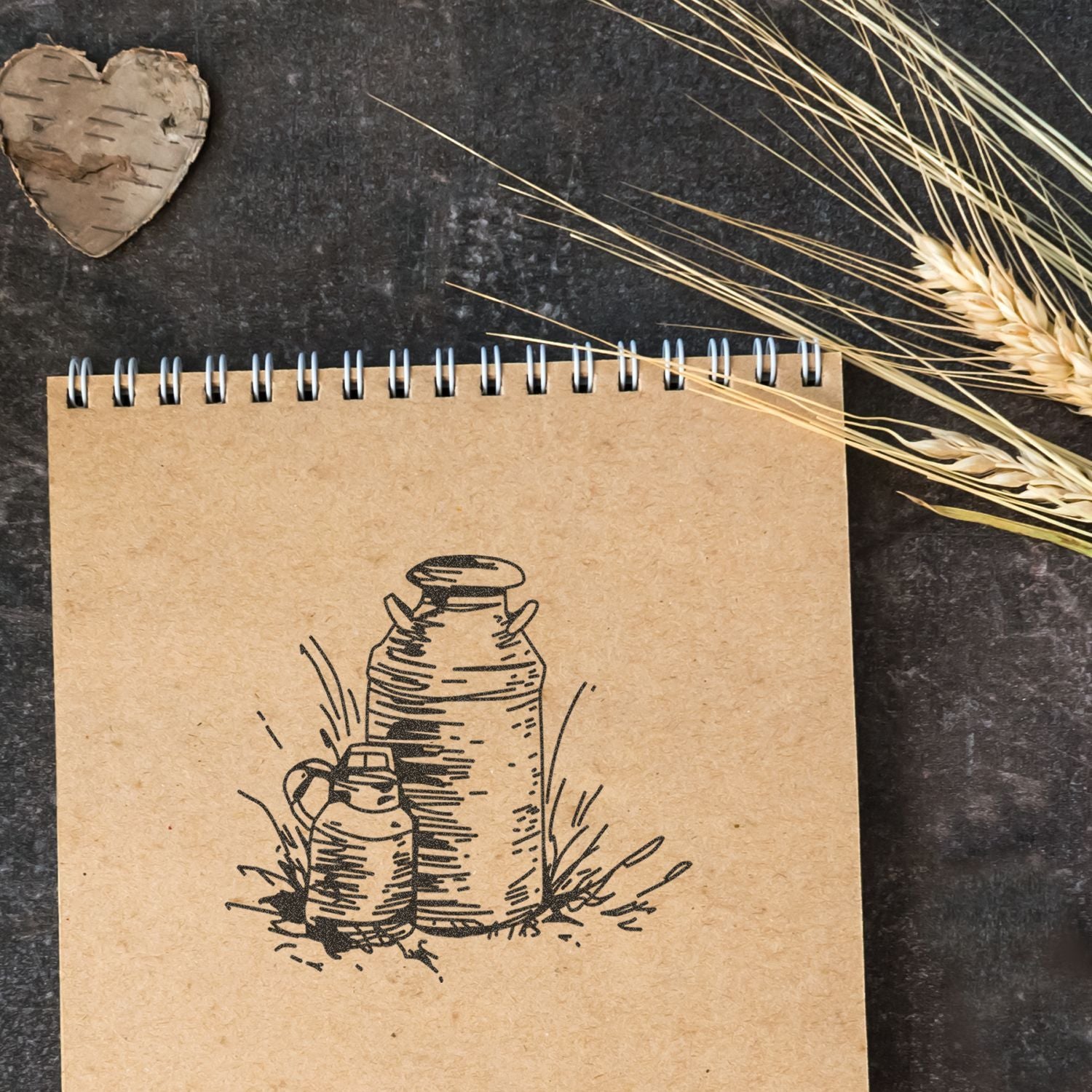 Rustic kraft notebook with vintage milk jug illustration, dried wheat, and birch heart on dark background.