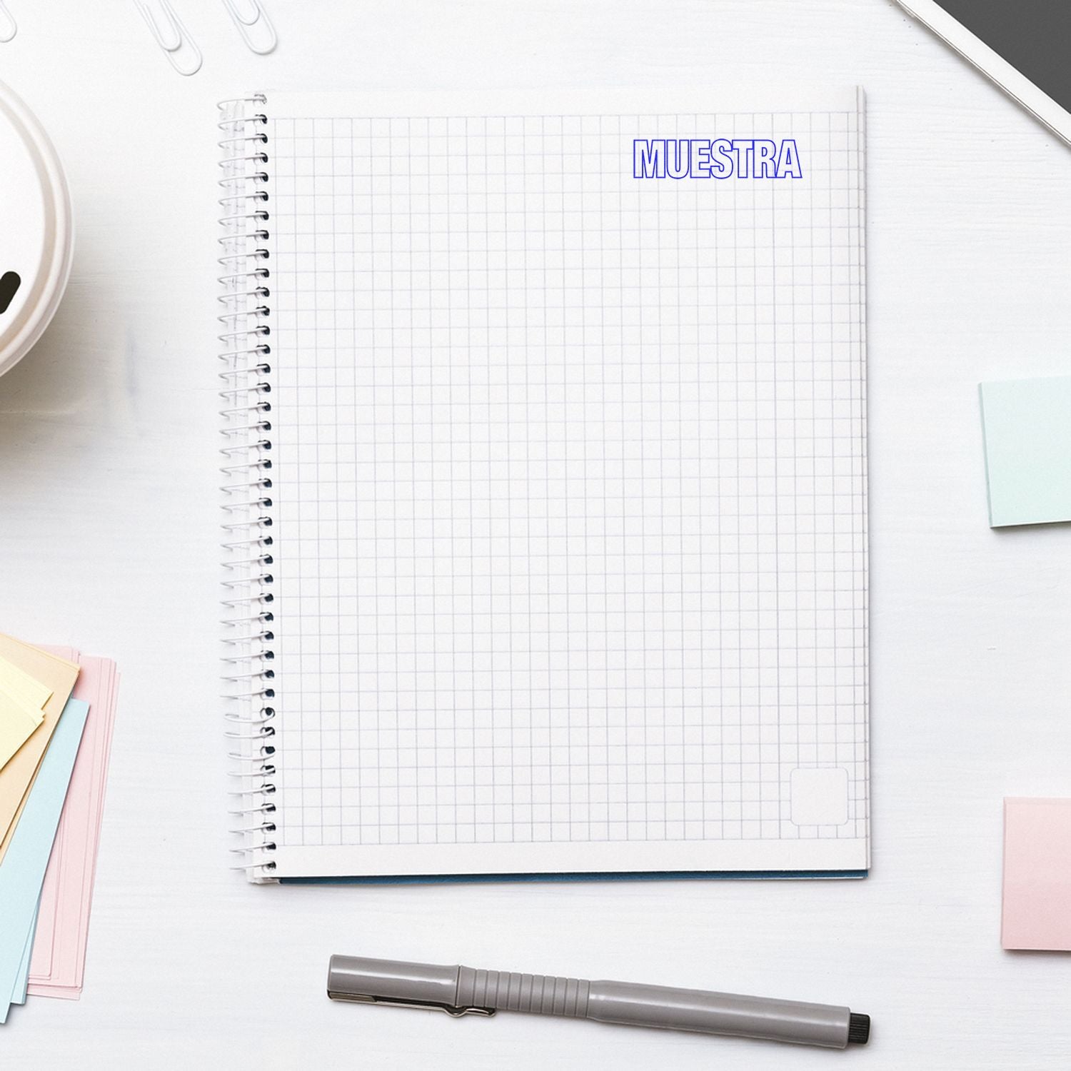 Cuaderno cuadriculado con marcador MUESTRA y material de oficina, ideal para plantillas y ejemplos de estudio o trabajo.