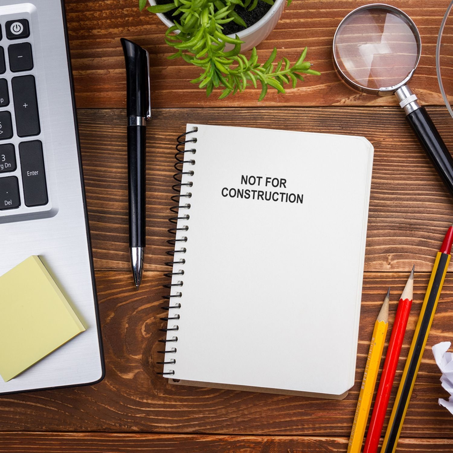 Notebook labeled Not for Construction on a wooden desk with laptop, pen, magnifying glass, pencils, and plant top view.
