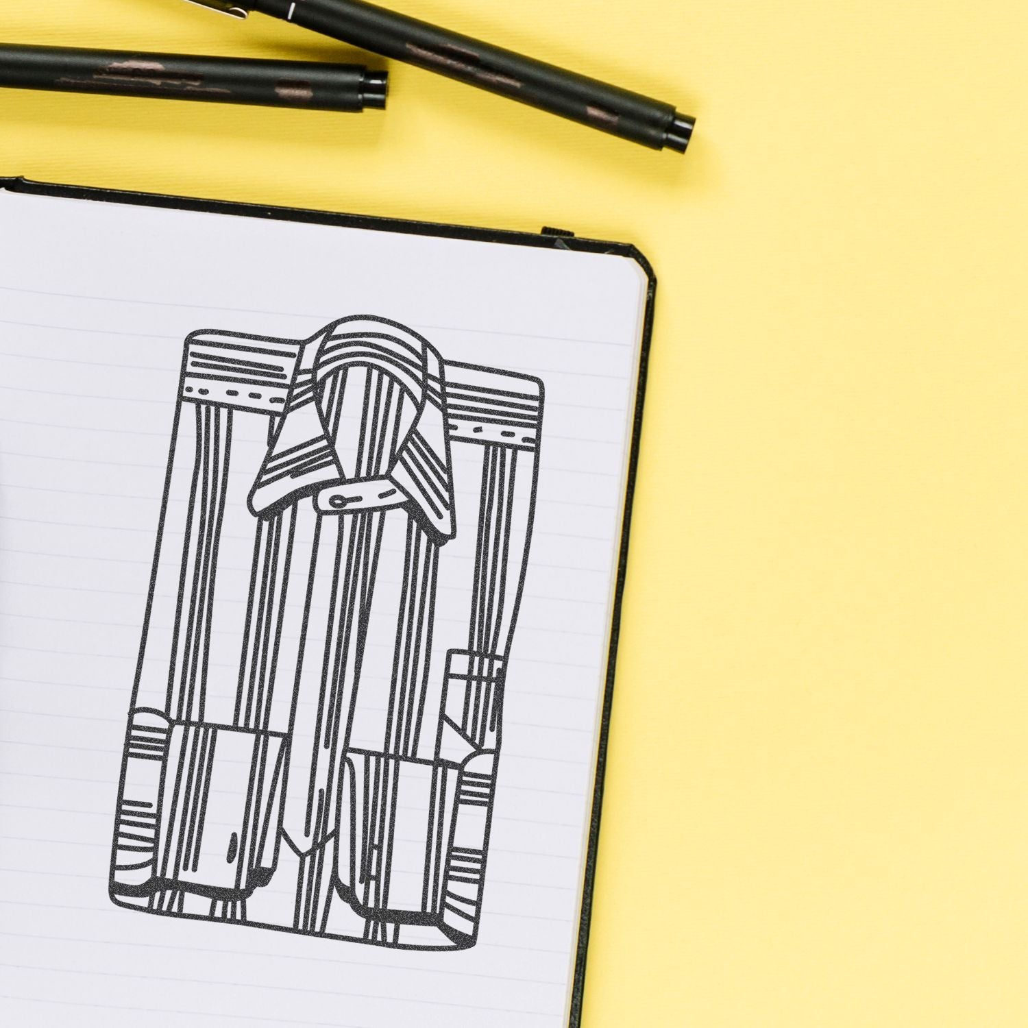 Fashion design sketch in a notebook on a yellow background with black pens.