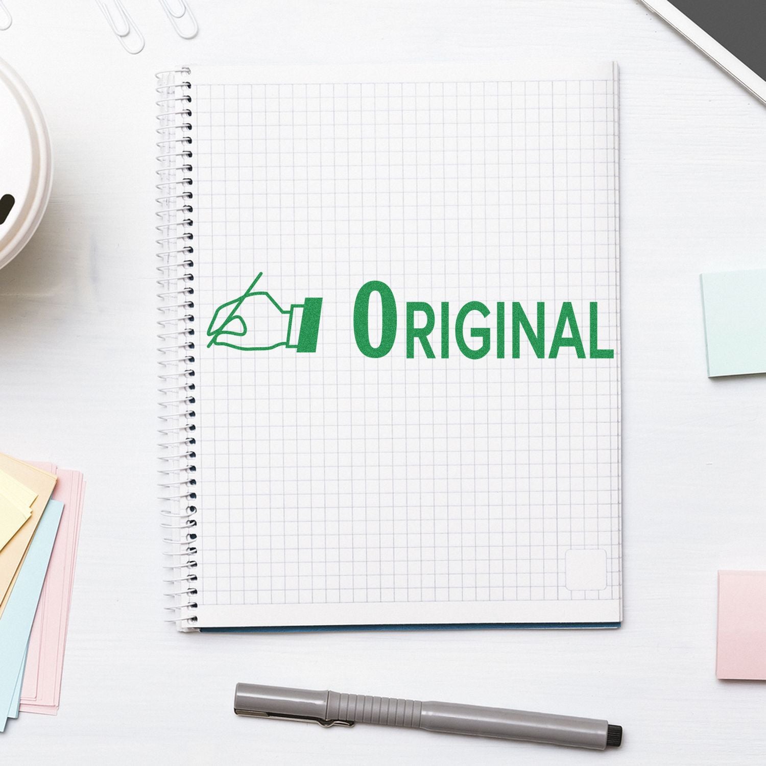 Top view of a spiral graph notebook with green Original logo on a clean white desk with pen and sticky notes.