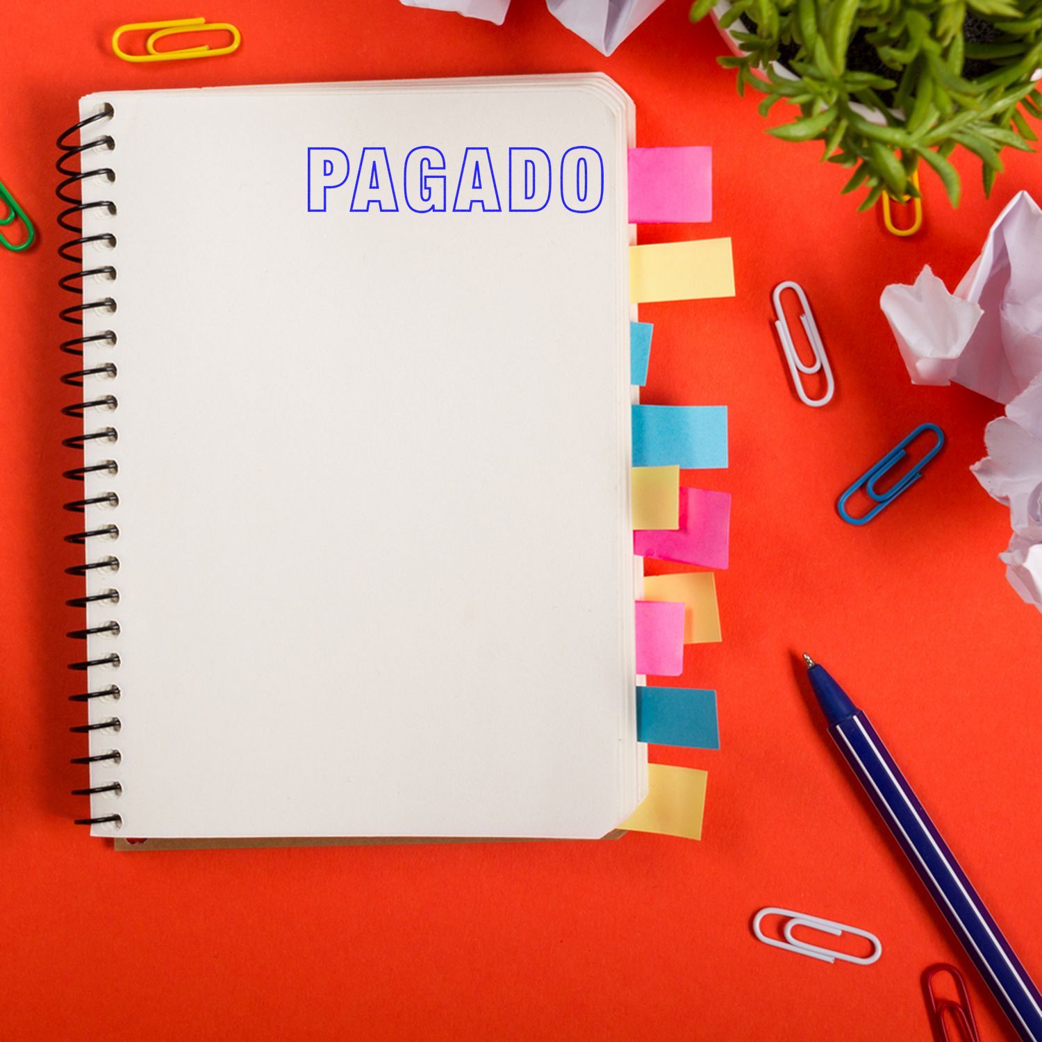 Cuaderno espiral con sello PAGADO, notas adhesivas, bolgrafo y clips de oficina sobre fondo rojo.