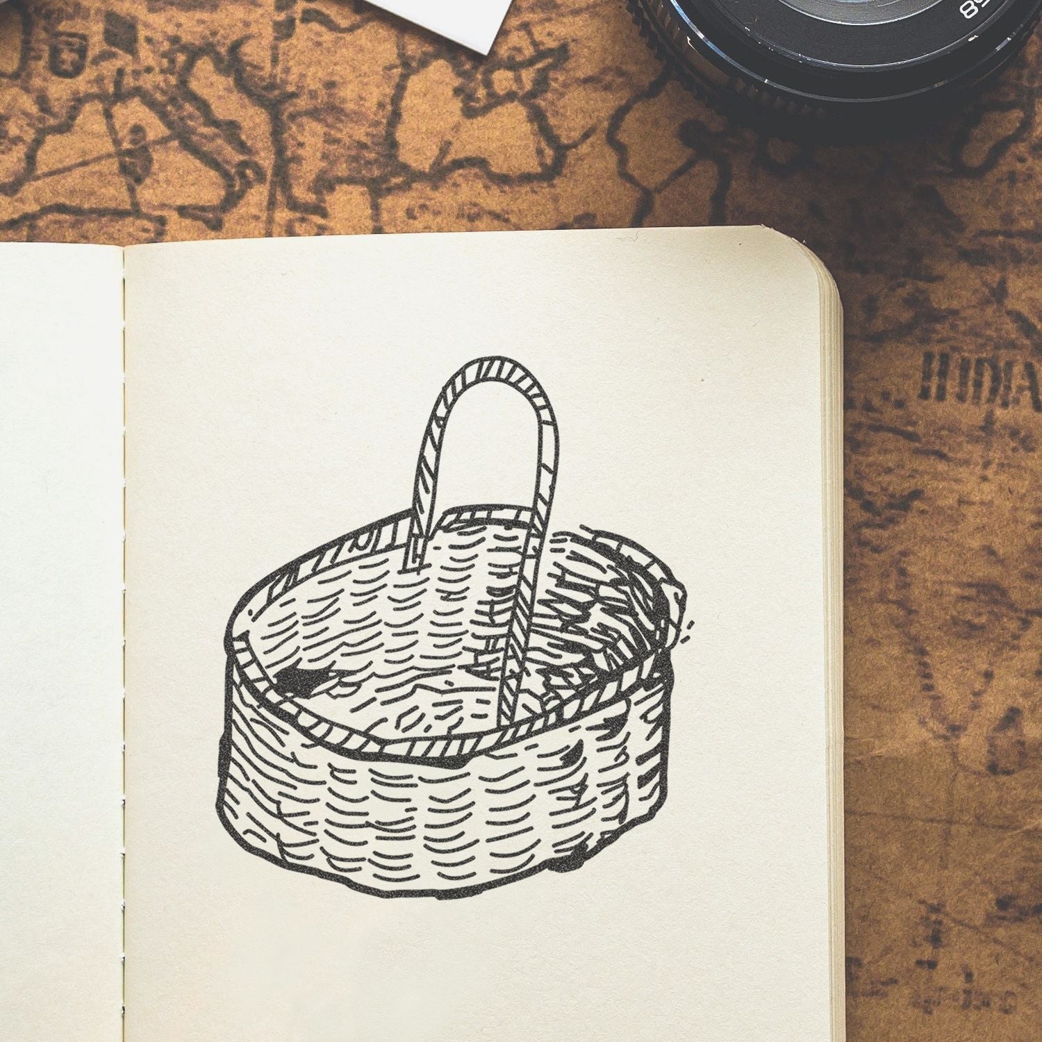 Open sketchbook with a black line drawing of a woven basket on a vintage map-style background, with a camera lens in the corner.