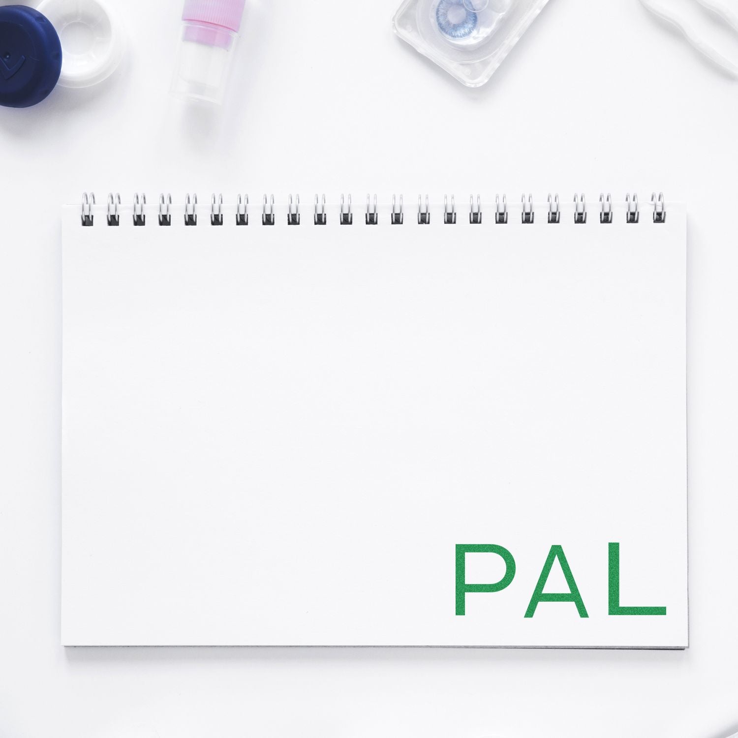 White spiral notebook labeled PAL with contact-lens case and solution bottles, ideal for tracking eye-care routines on a clean desk.