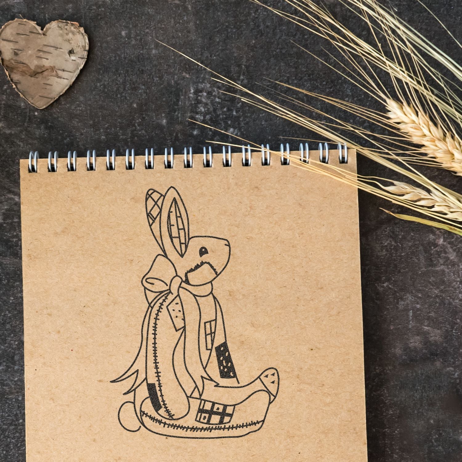 Rustic spiral notebook with cute abstract bunny illustration on kraft paper, styled with dried wheat on a dark background.