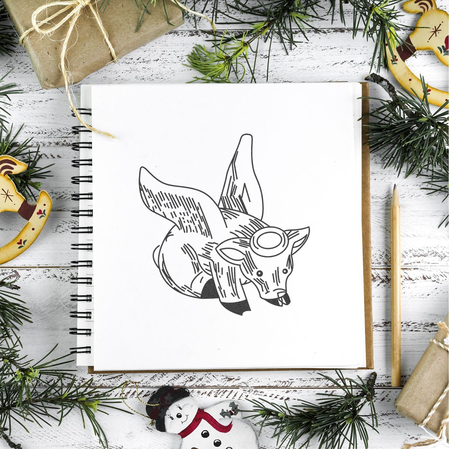 Holiday craft scene with a sketchbook displaying a fox illustration, surrounded by pine branches, wrapped gifts, and Christmas decorations on a rustic white wooden table.