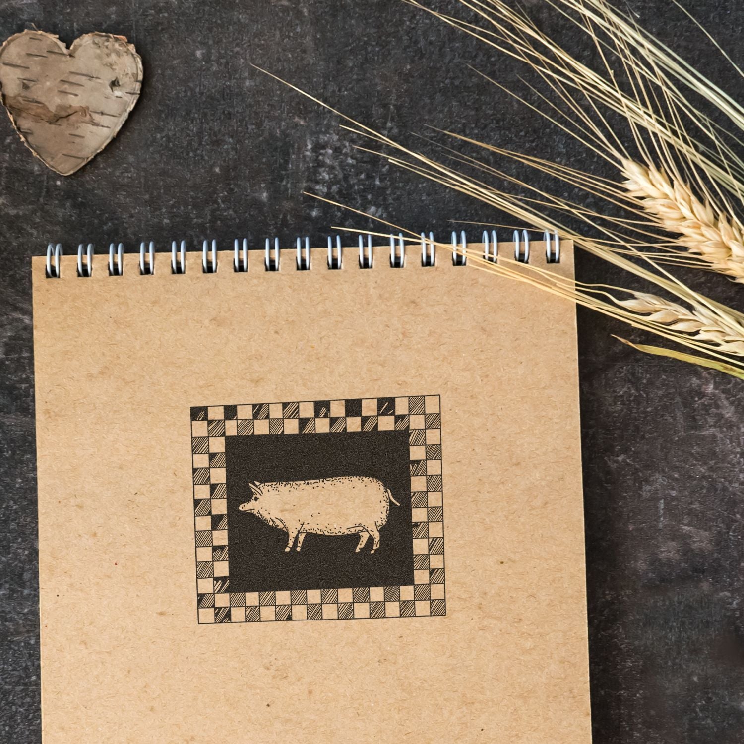Rustic kraft paper spiral notebook with pig illustration, wheat stalks, and wooden heart on a dark background