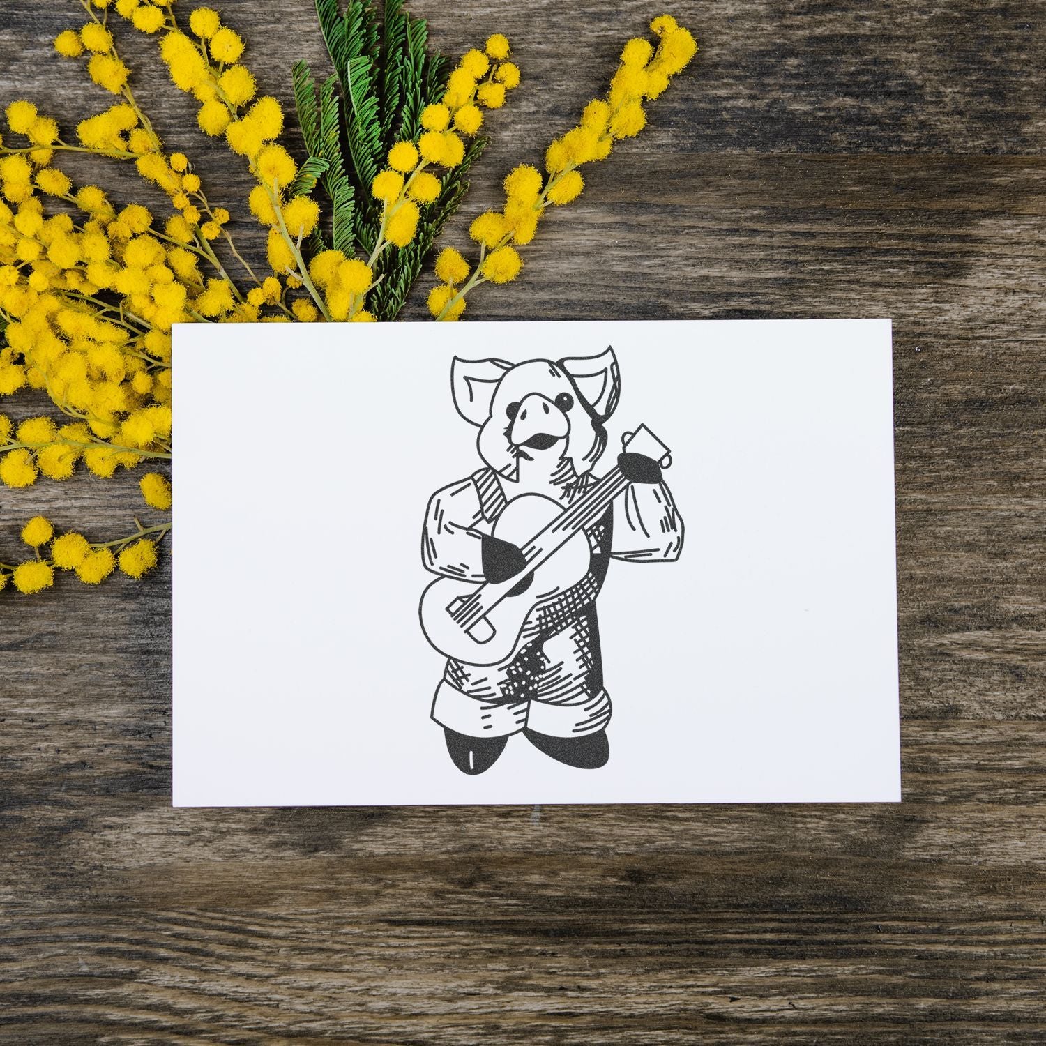 Handmade card with pig playing guitar illustration on rustic wood table with yellow mimosa flowers.