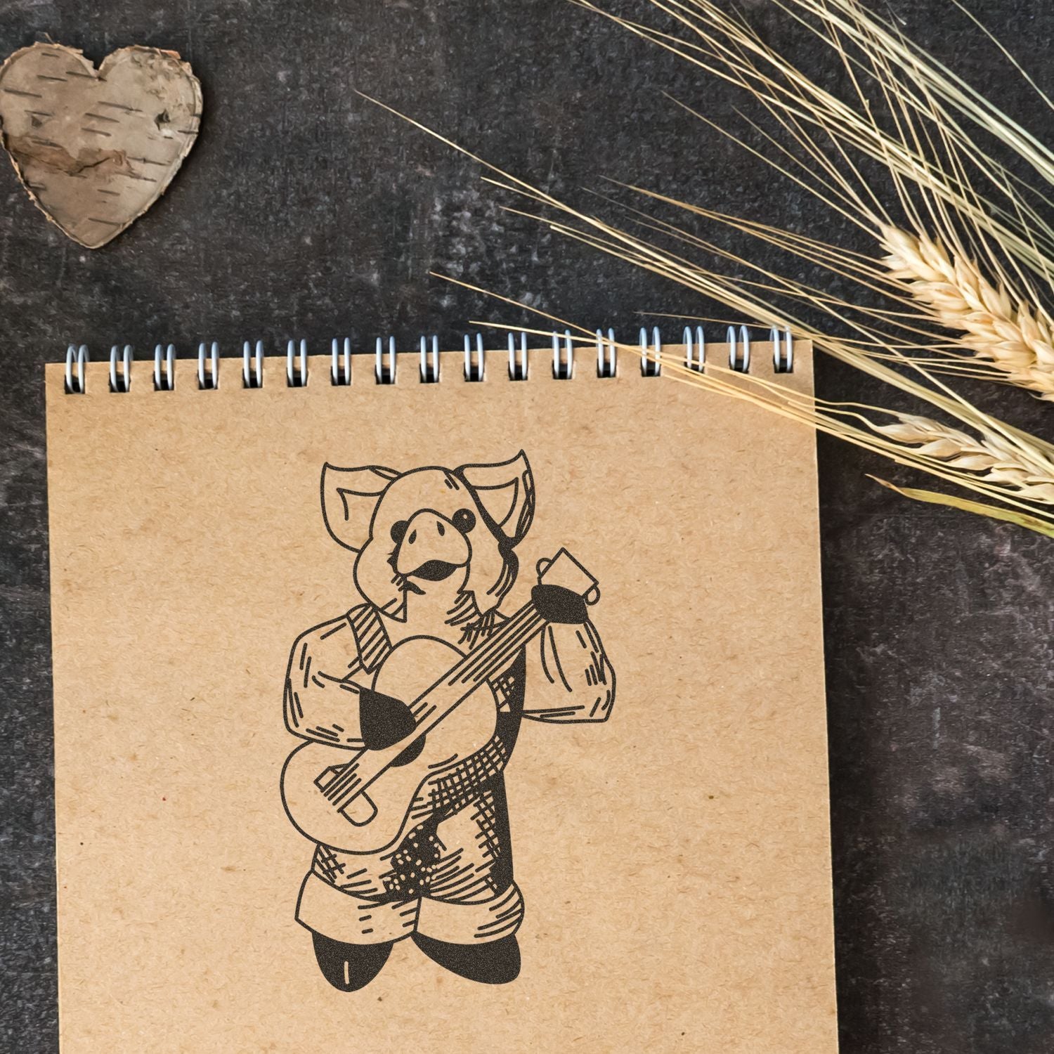 Rustic spiral notebook with a pig playing guitar illustration, styled with dried wheat and a wooden heart on a dark background.