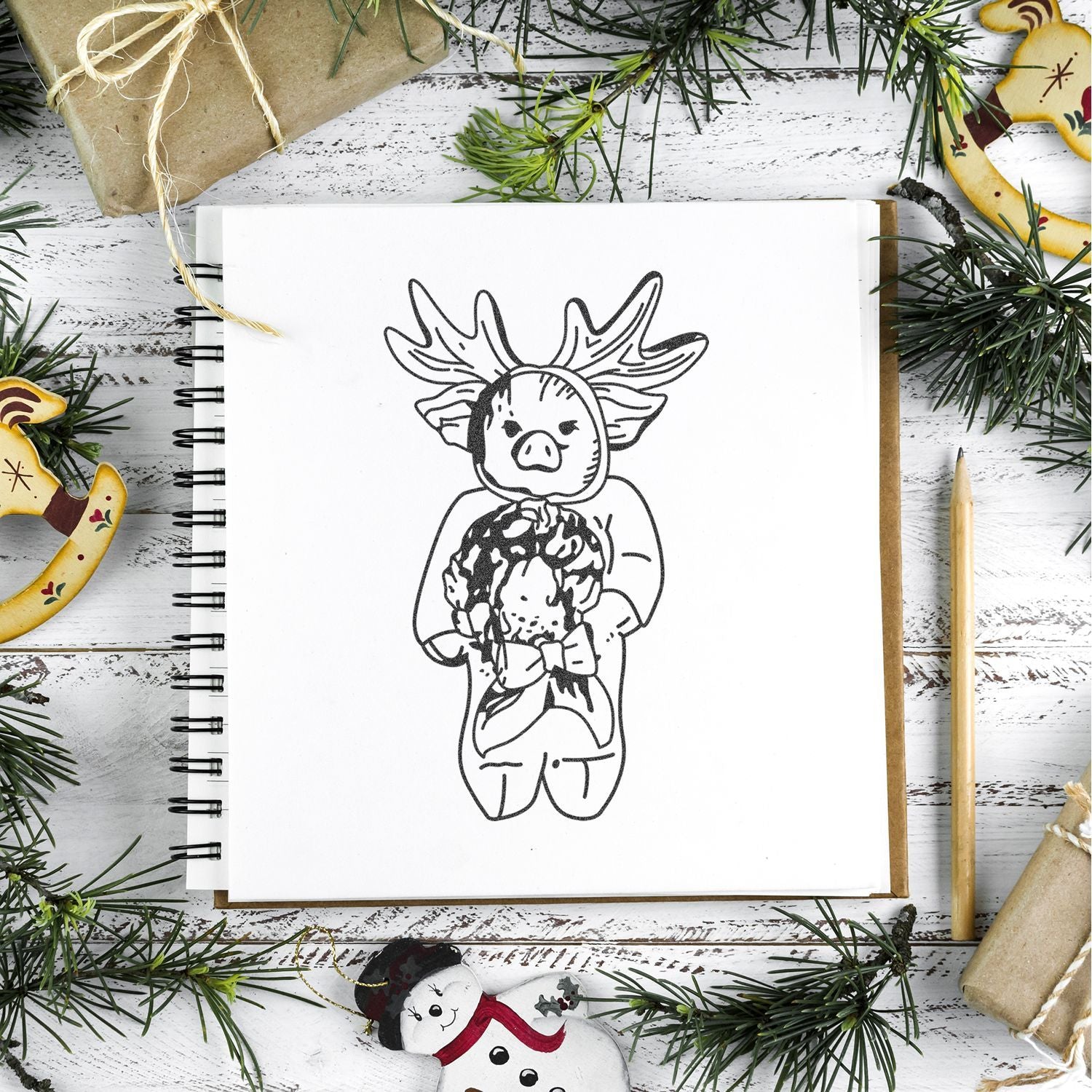 Christmas-themed sketchbook with cute hand-drawn reindeer pig illustration, surrounded by pine branches, wrapped gifts, and holiday ornaments on a rustic white wooden background