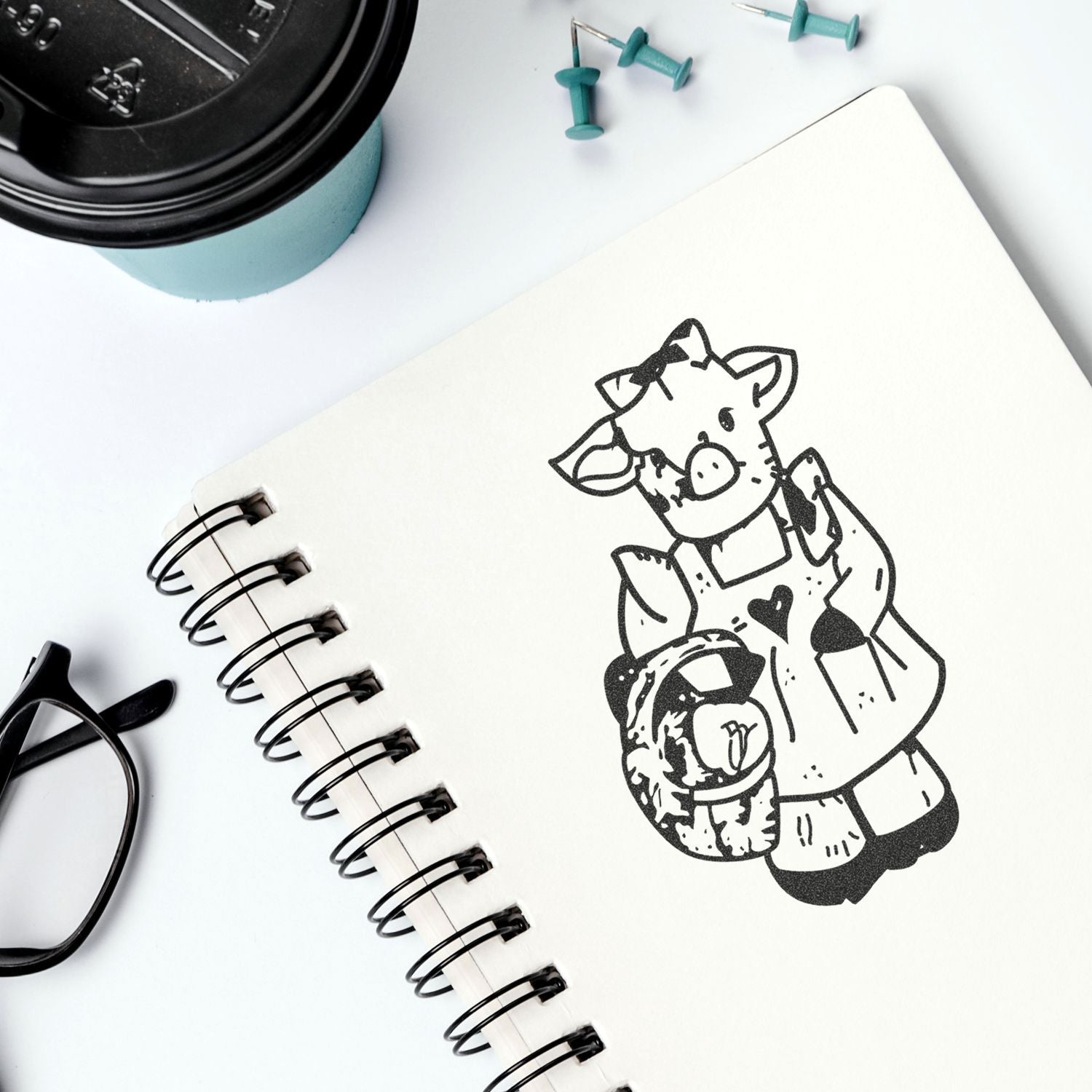 Spiral notebook with cute pig illustration on a white desk, alongside a coffee cup, push pins, and glasses, ideal for stationery and cute notebook design concepts