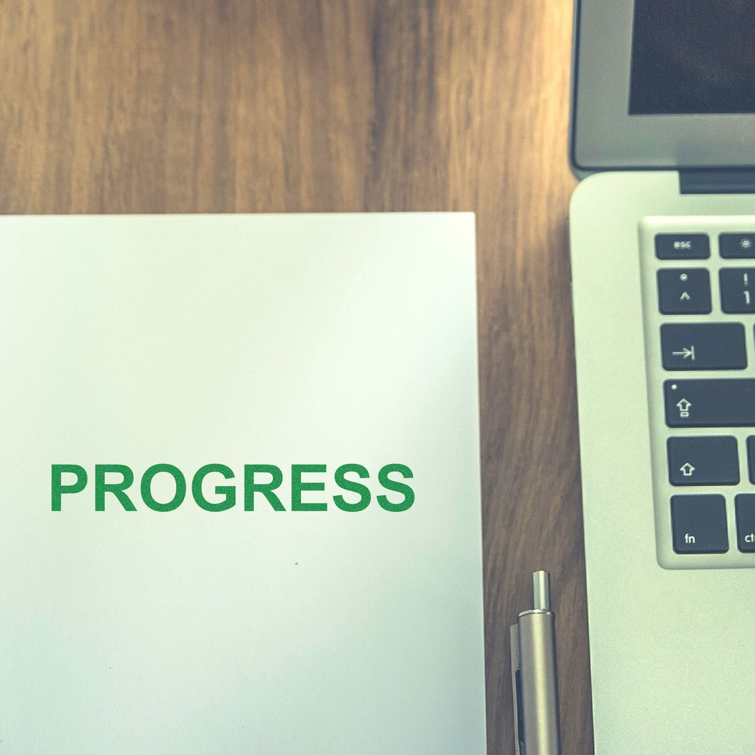 Green PROGRESS text on a notebook beside a laptop and pen on a wooden desk, symbolizing productivity and business growth.
