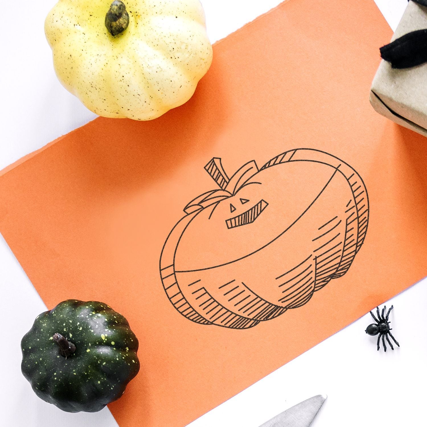 Halloween pumpkin illustration on orange paper surrounded by decorative gourds, a spider, and craft supplies