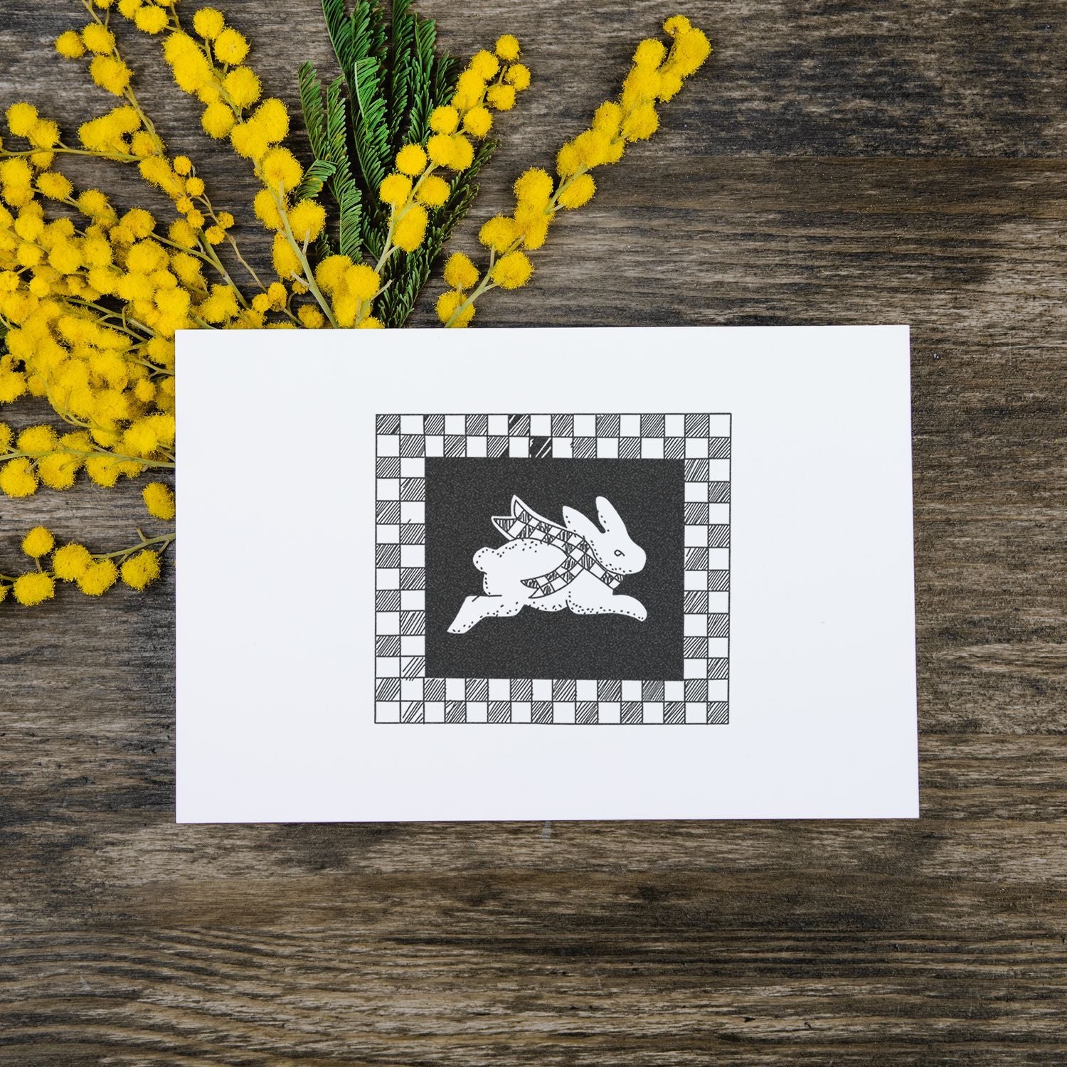 White greeting card with black-and-white rabbit illustration surrounded by yellow mimosa flowers on a rustic wooden table.