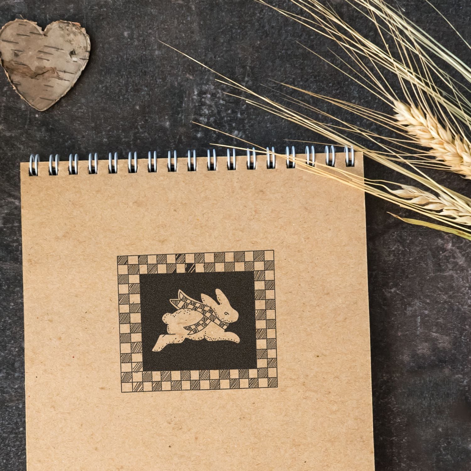 Rustic kraft paper spiral notebook with bunny illustration, dried wheat, and wooden heart on dark background.