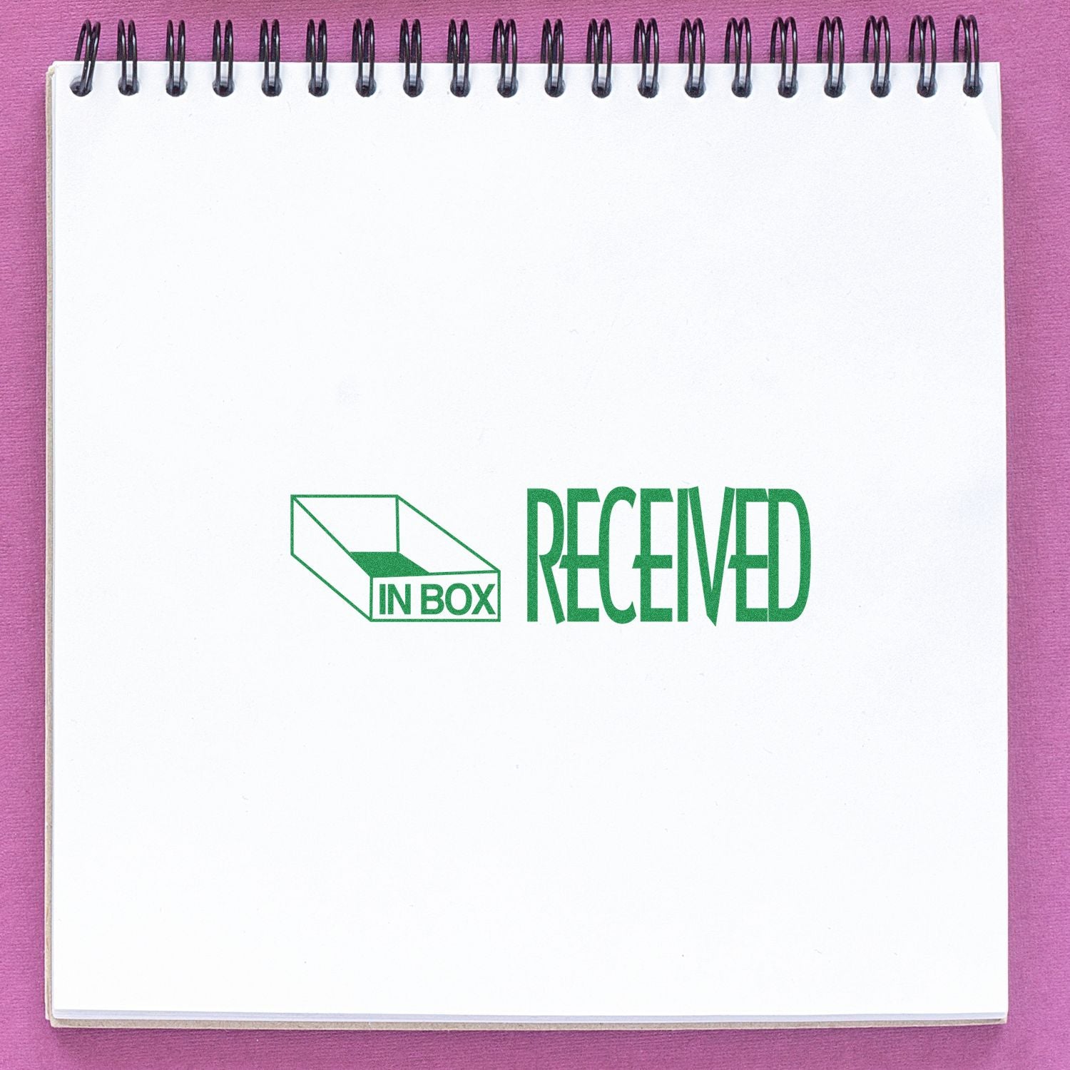 Spiral notebook with In Box Received green logo on a pink background, minimal office and packaging concept.