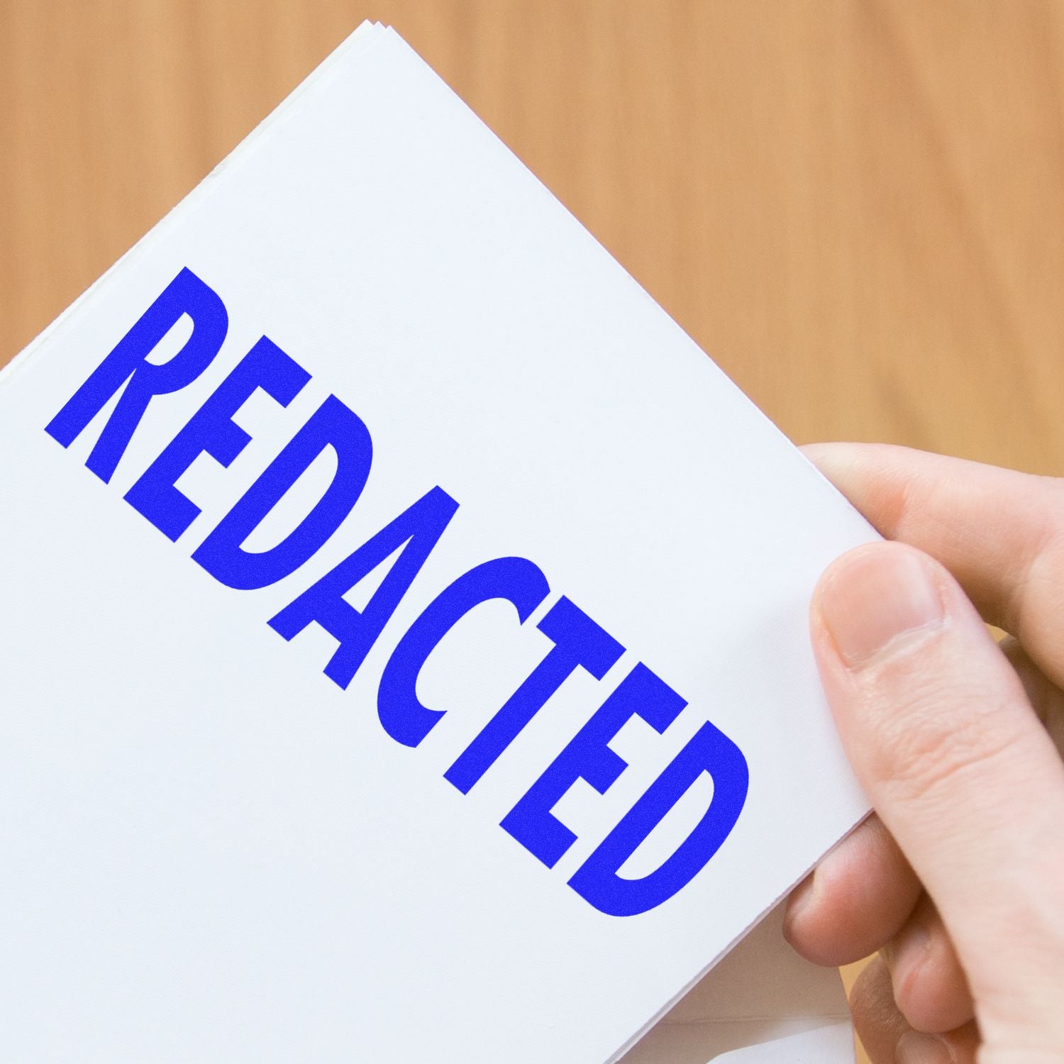Hand holding a white document stamped with bold blue REDACTED text on a wooden background.