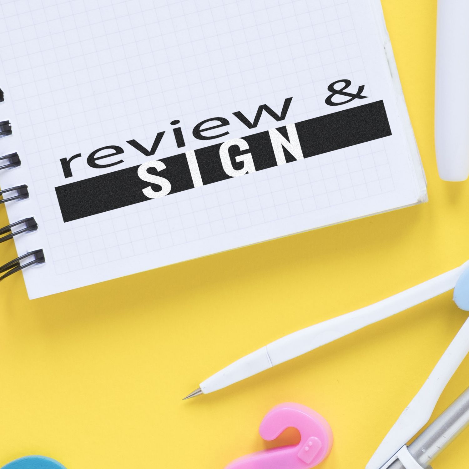 Notebook with review & sign text, pens, and office supplies on a yellow background, concept for document approval and signing.