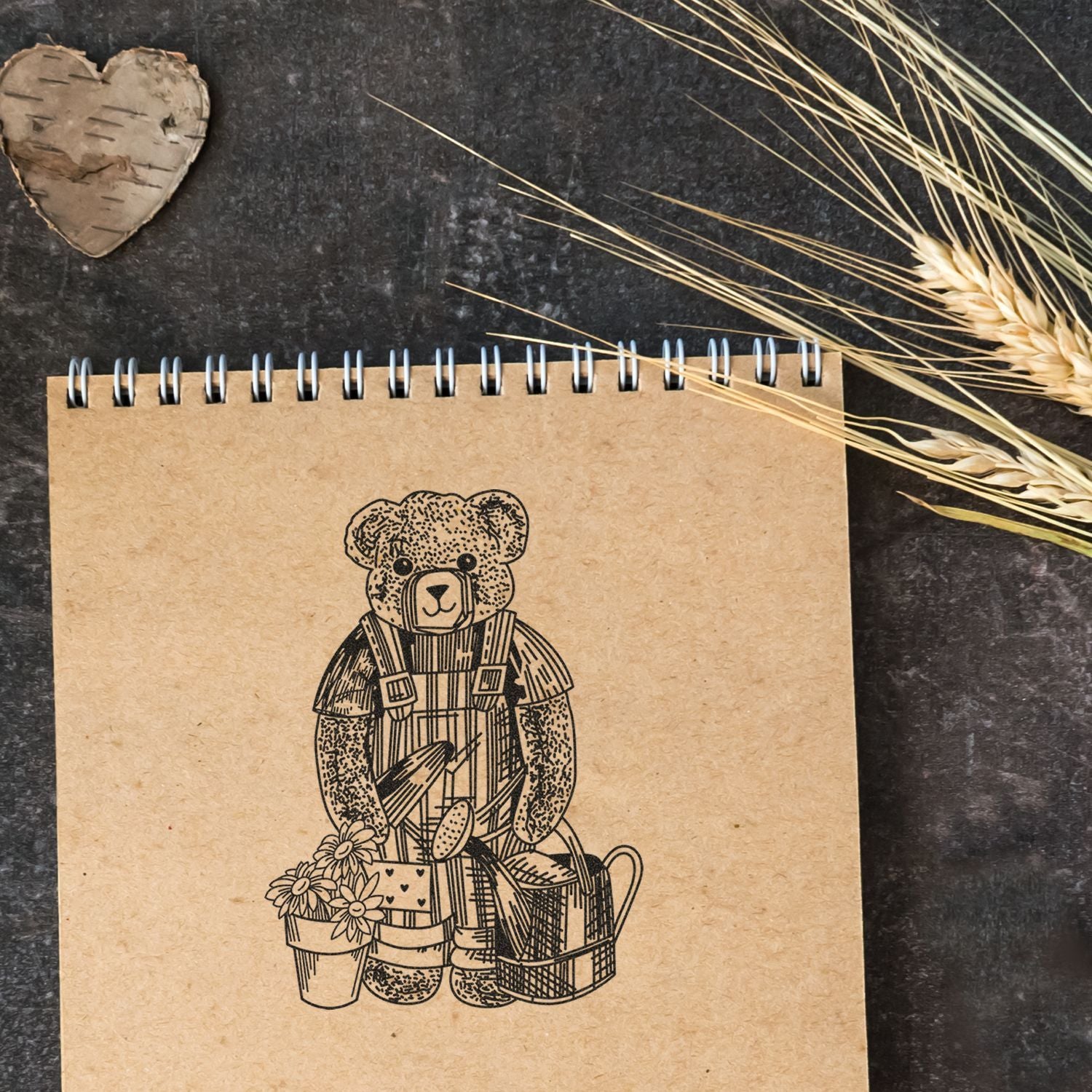 Kraft spiral notebook with teddy bear illustration, rustic dried wheat decor, and wooden heart ornament on a dark background.