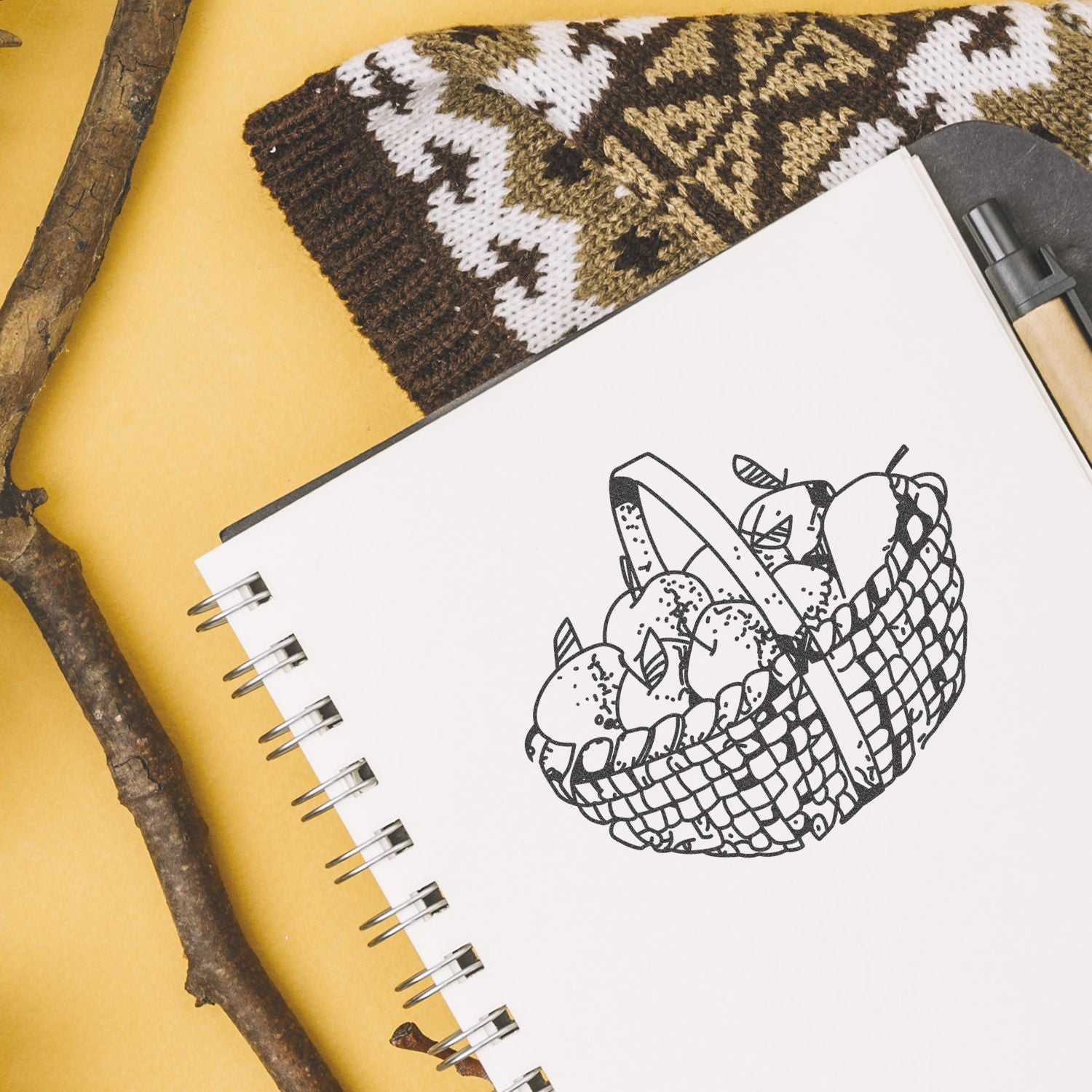 Flat lay of a sketchbook with a hand-drawn fruit basket illustration, pen, woven textile, and rustic sticks on a yellow background.