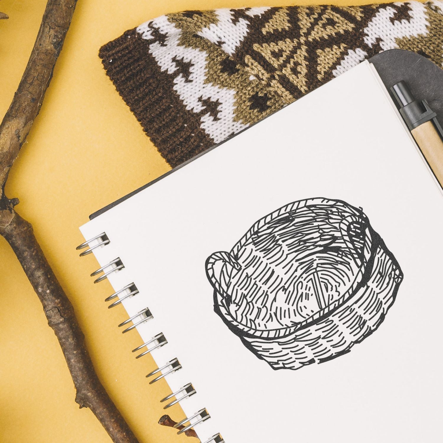 Spiral sketchbook with a hand-drawn woven basket illustration on a yellow background, styled with a branch and patterned textile.