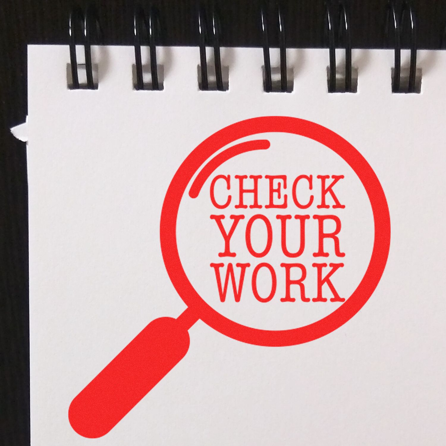 Red magnifying glass icon with the words Check Your Work on a spiral notebook page, symbolizing proofreading and quality review.