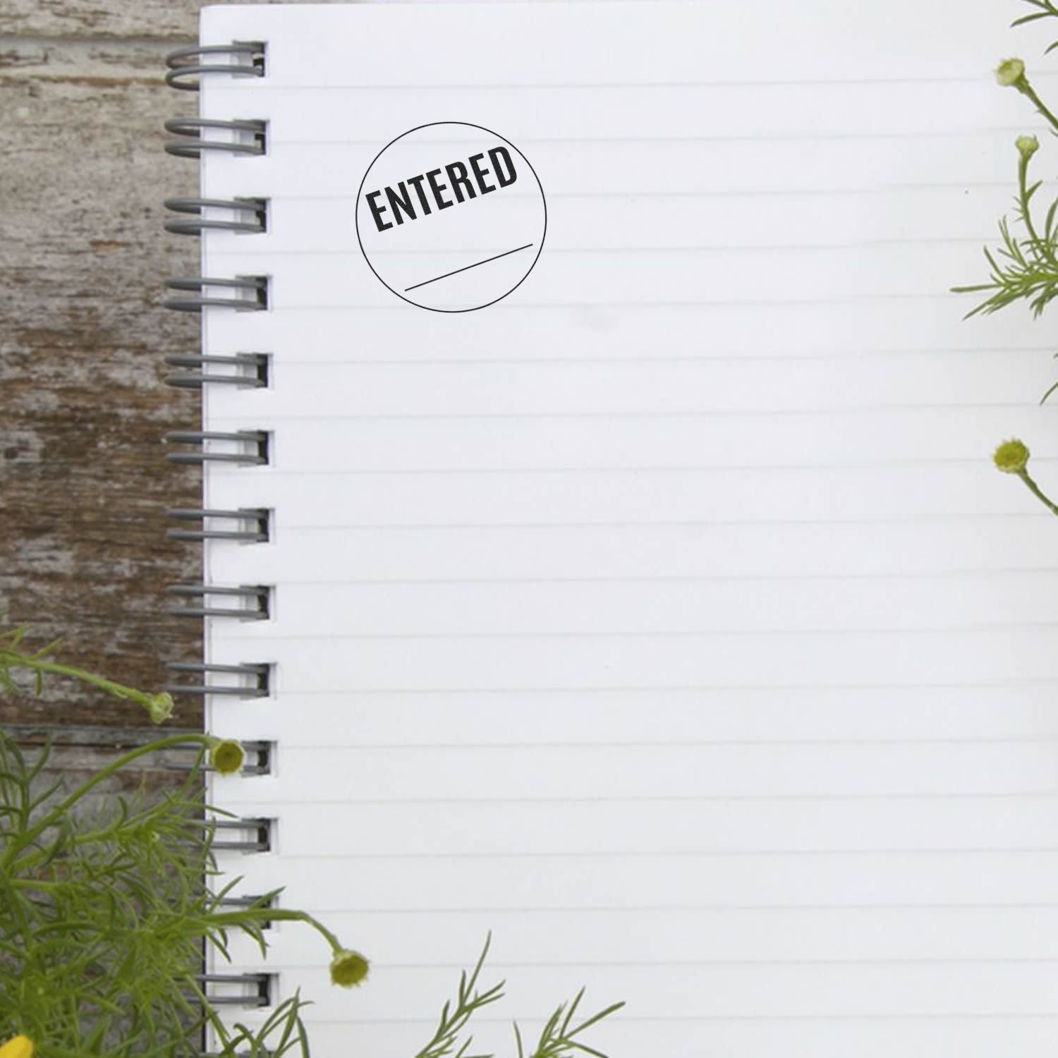 Blank spiral notebook with an Entered stamp on a rustic wooden table, surrounded by small flowers.