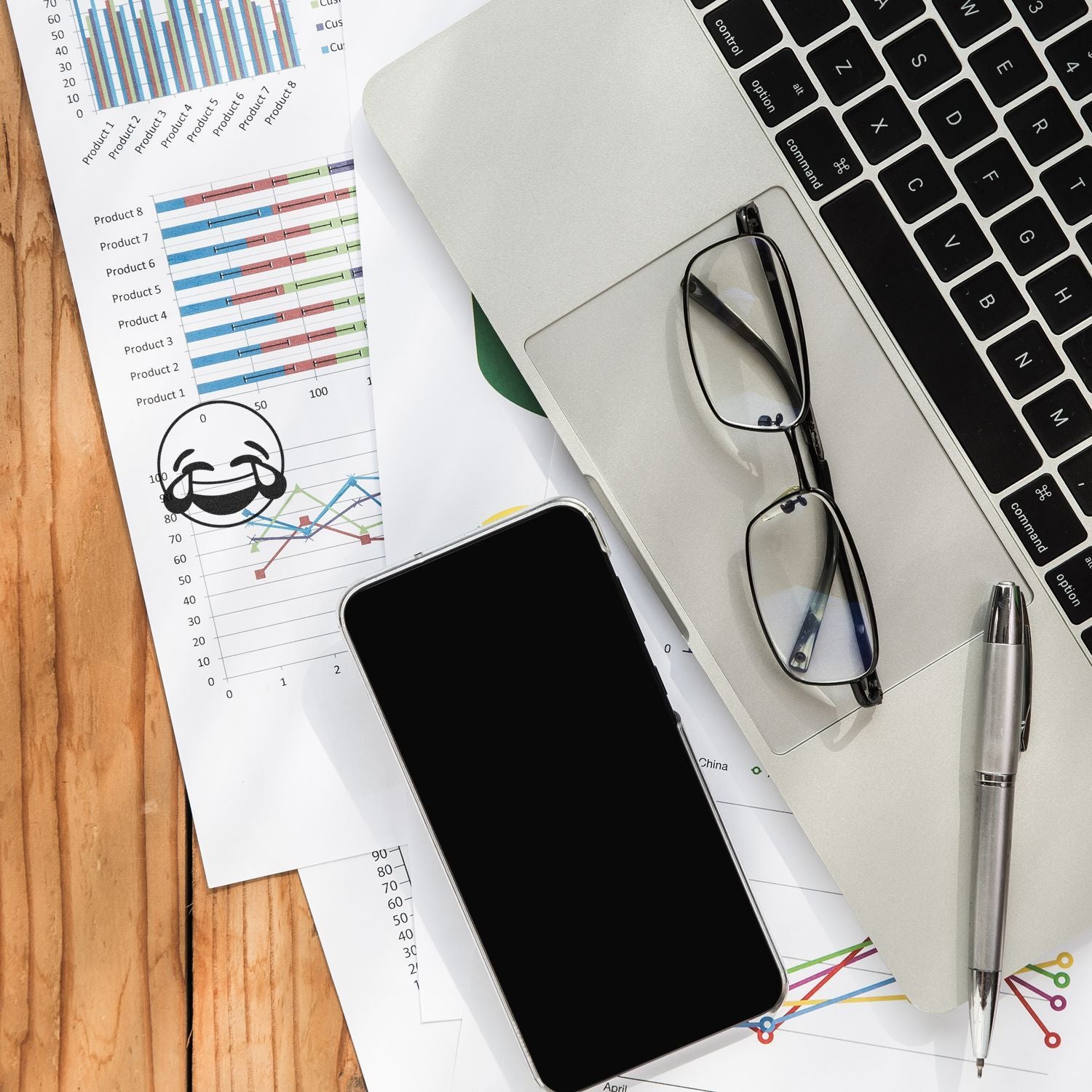 Office desk with laptop, smartphone, eyeglasses, pen, and business analytics charts on wooden table