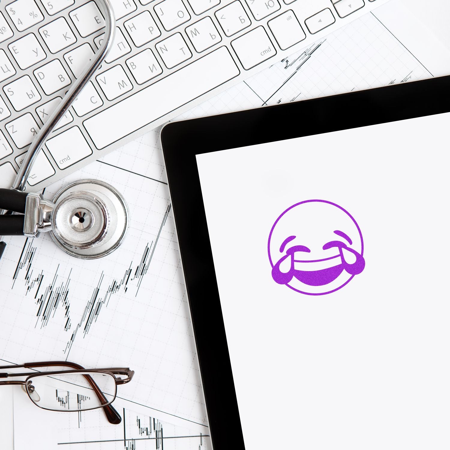 Tablet with laughing emoji on screen, stethoscope, eyeglasses, and stock market chart on a laptop keyboard background