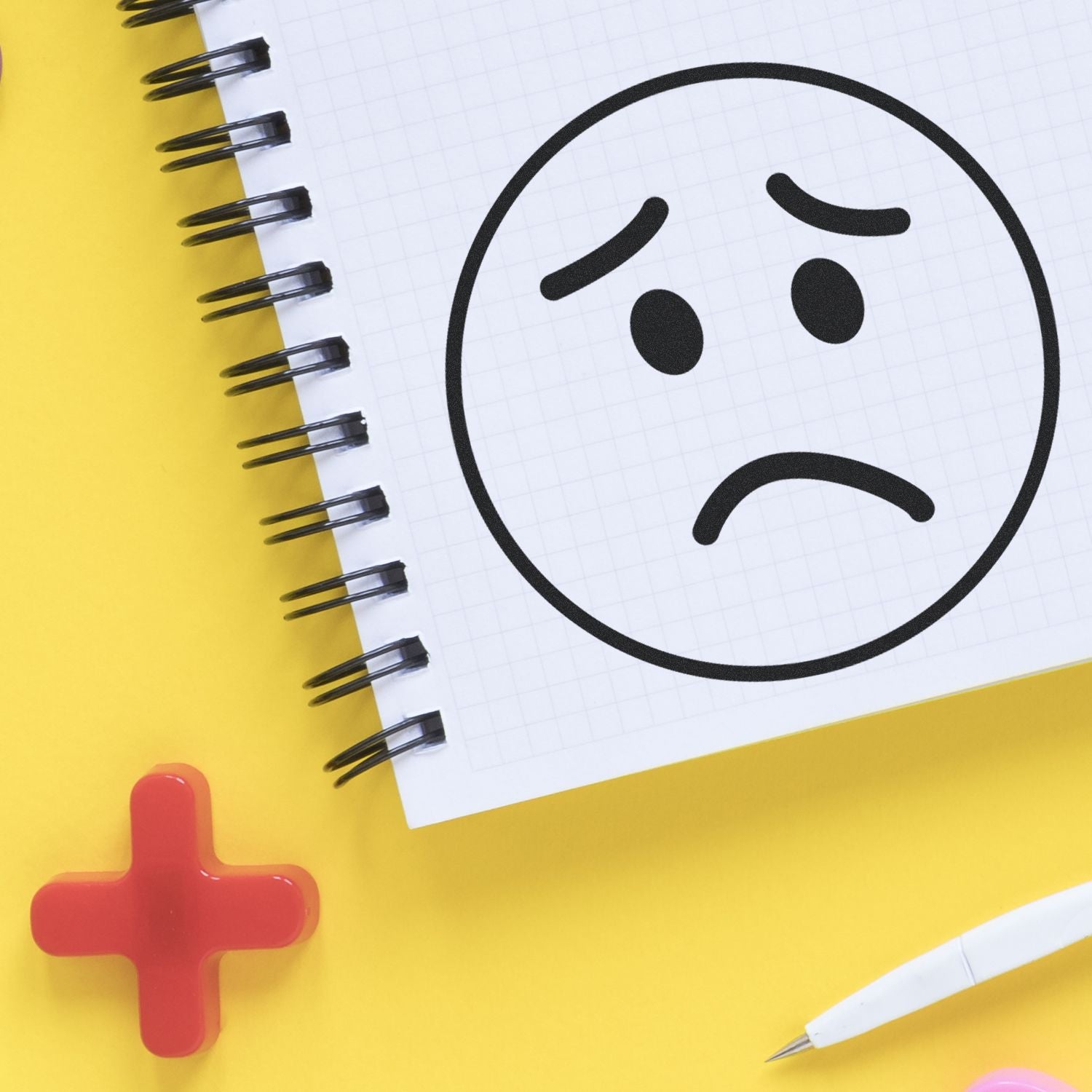 Sad face doodle in a spiral notebook on a yellow background with red plus sign and pen