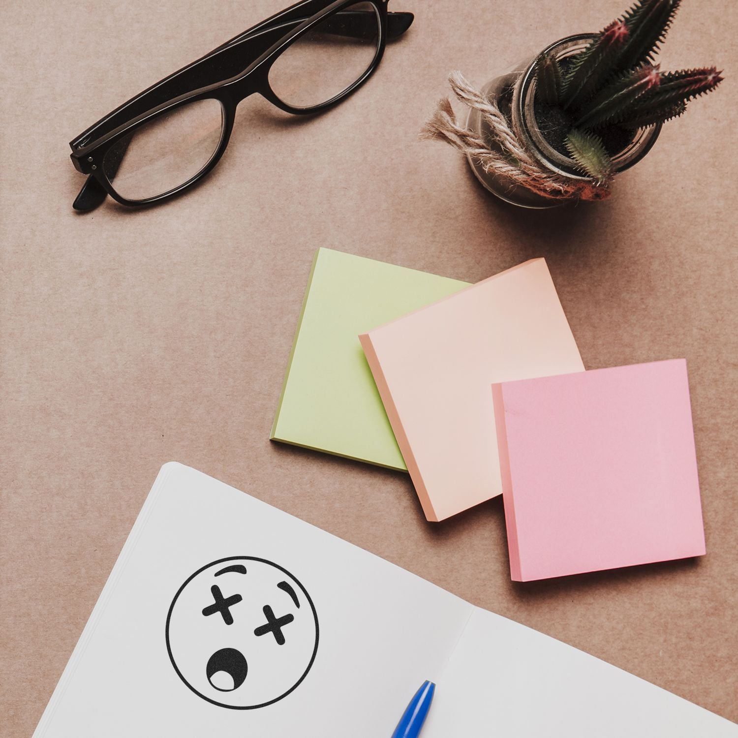 Flat lay of creative office desk with black eyeglasses, pastel sticky notes, notebook, pen, and small potted succulent plant on a brown background.