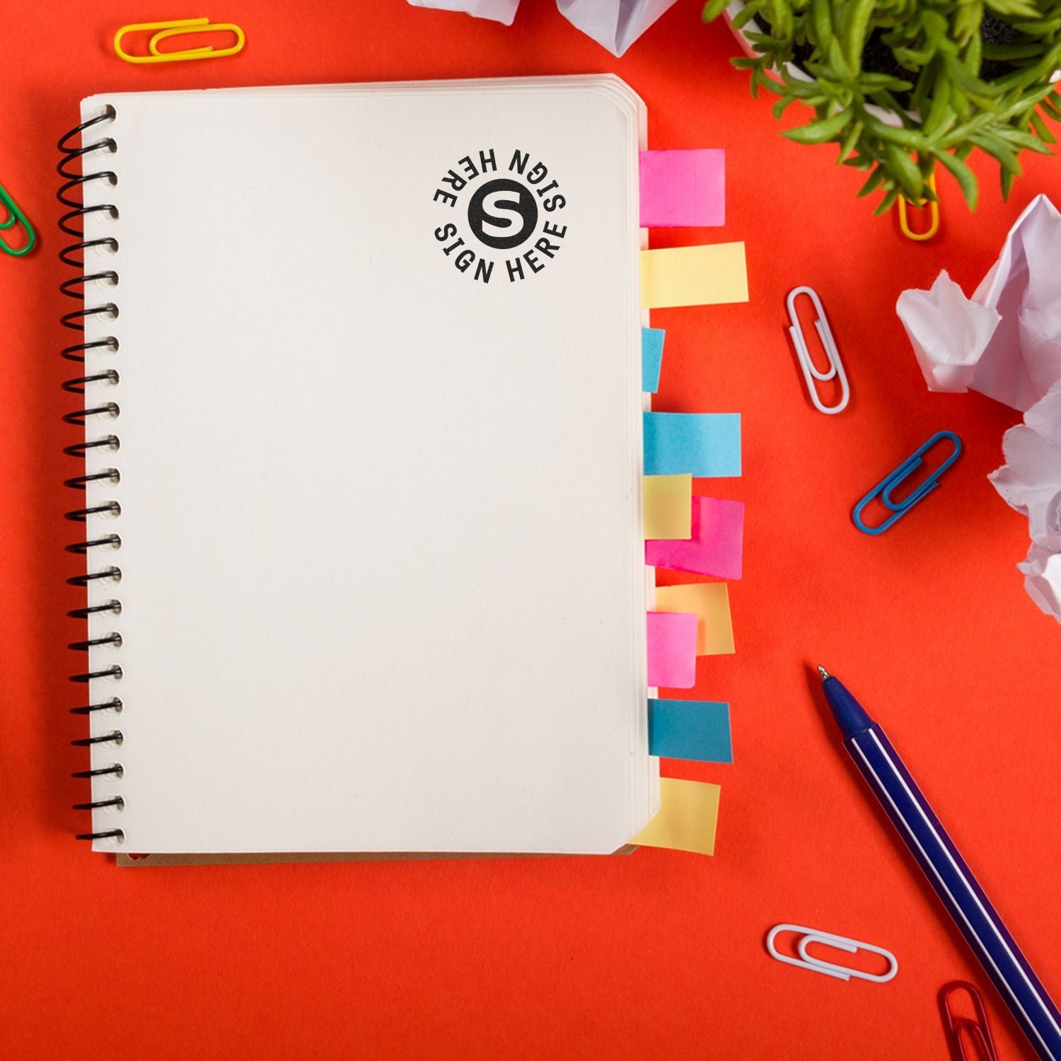 Blank spiral notebook with colorful sticky notes, pen, paper clips, and office supplies on an orange desk workspace.