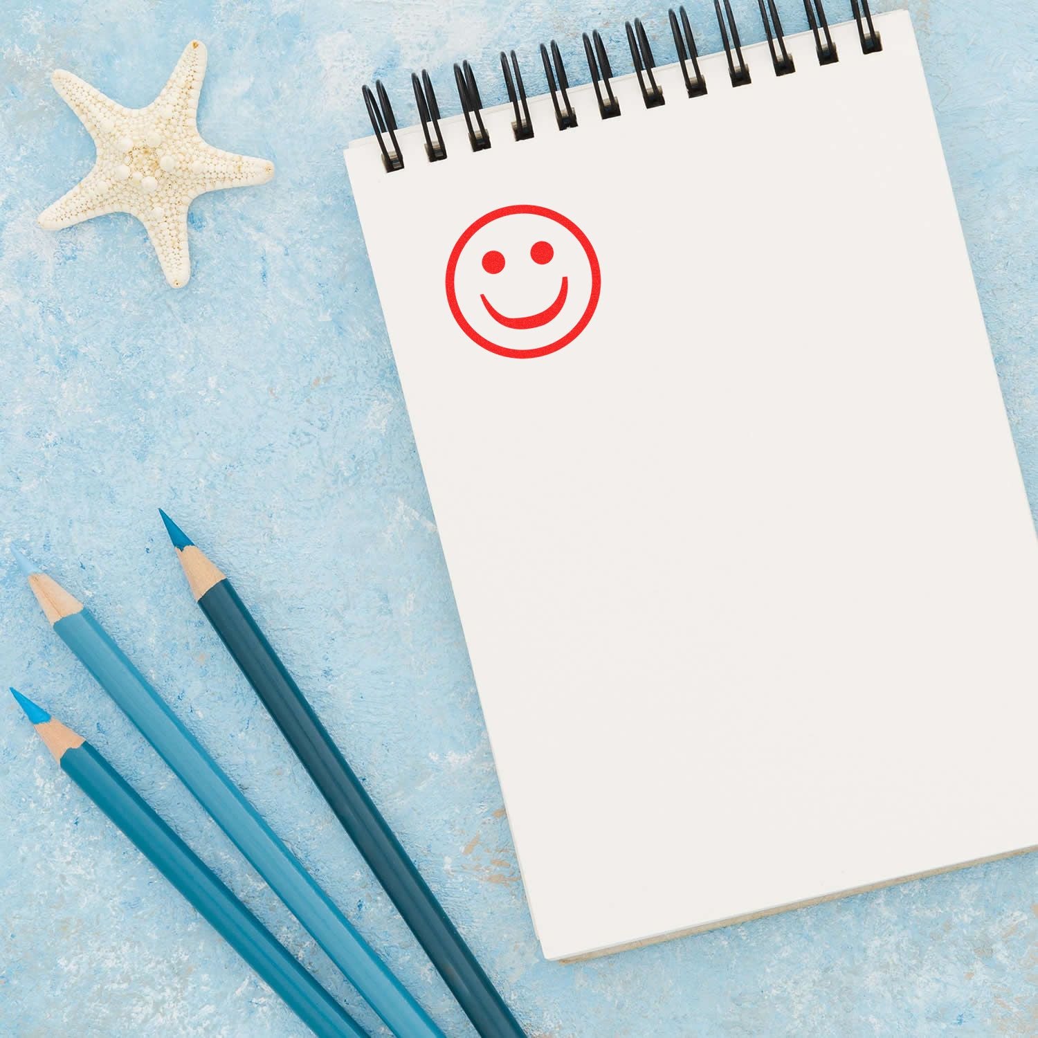 Blank spiral notebook with smiley face, blue pencils, and starfish on a summer beach-themed background.