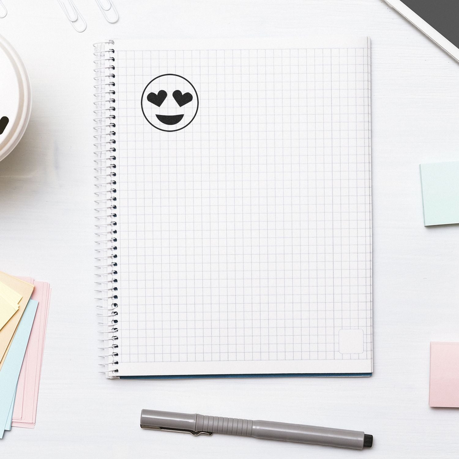 Top view of a spiral notebook with heart eyes emoji, pen, and pastel sticky notes on a clean white desk flat lay.