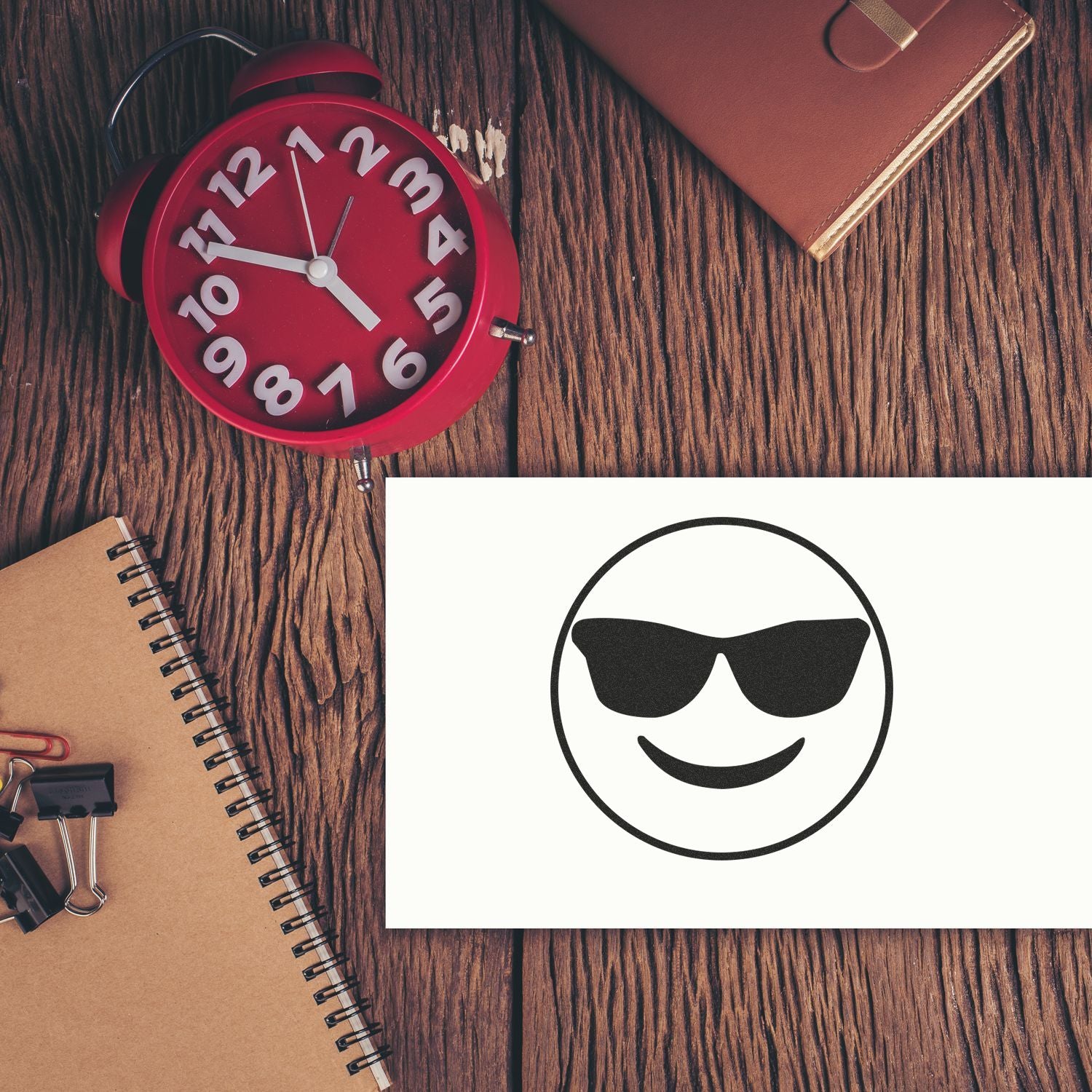 Red alarm clock, notebook, and sunglasses smiley on a rustic wooden desk, concept of morning, time management, and relaxed office lifestyle.
