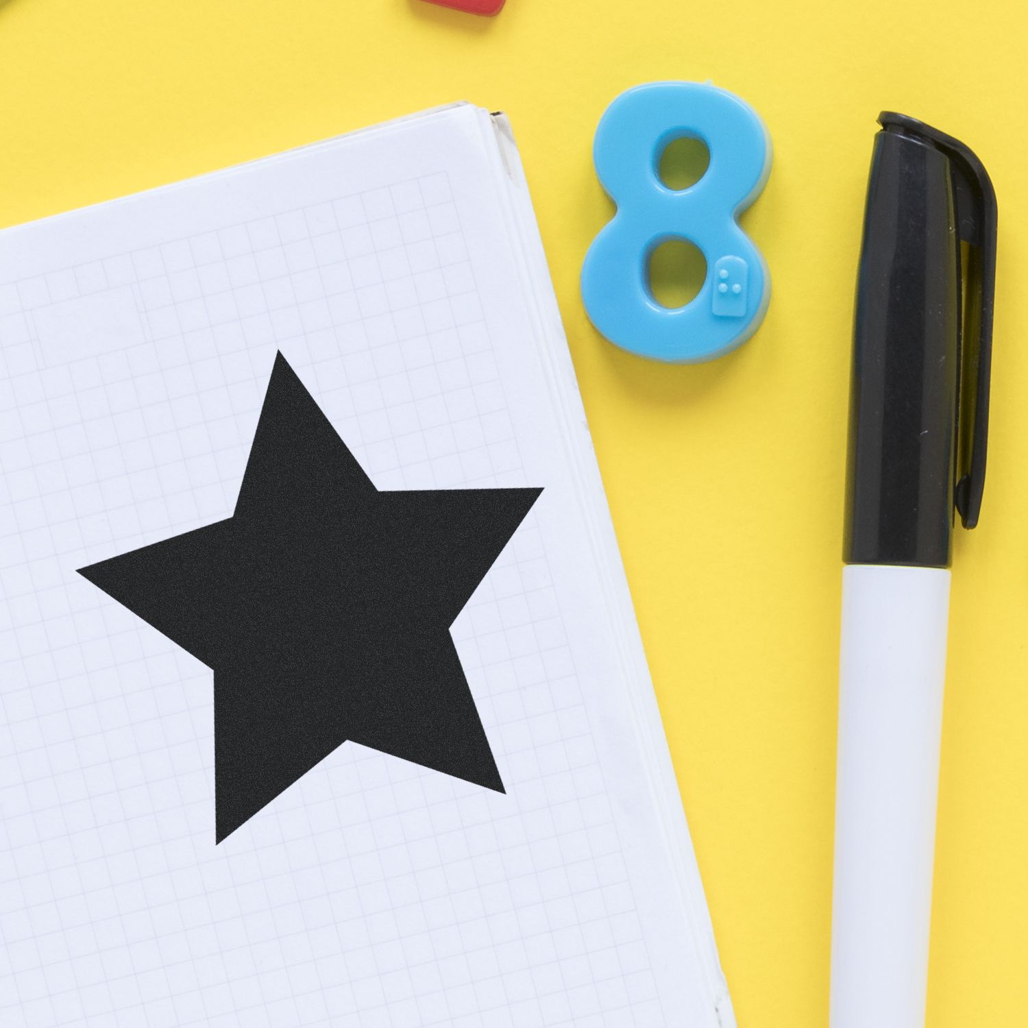 Black star on graph paper notebook with number 8 magnet and marker on yellow background, creative school supplies flat lay.