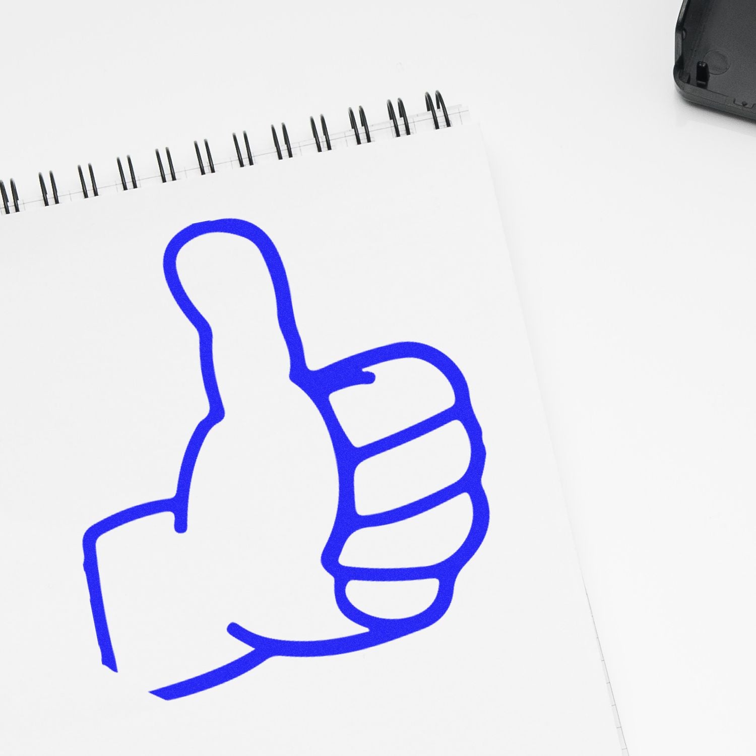 Blue thumbs up icon drawn on a spiral notebook on a white desk, symbolizing approval, positive feedback, and success.
