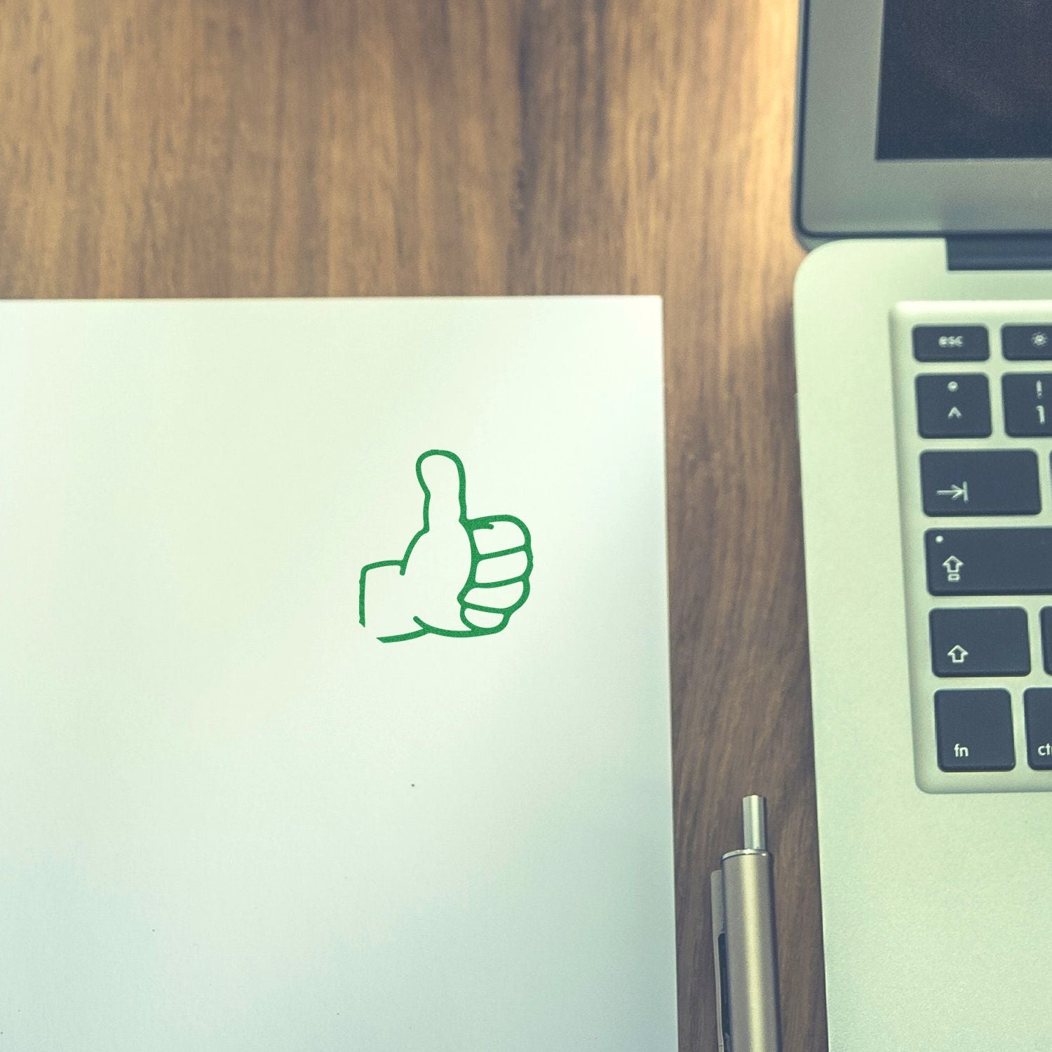 Green thumbs up icon on white paper next to a laptop on a wooden desk, top view office workspace.