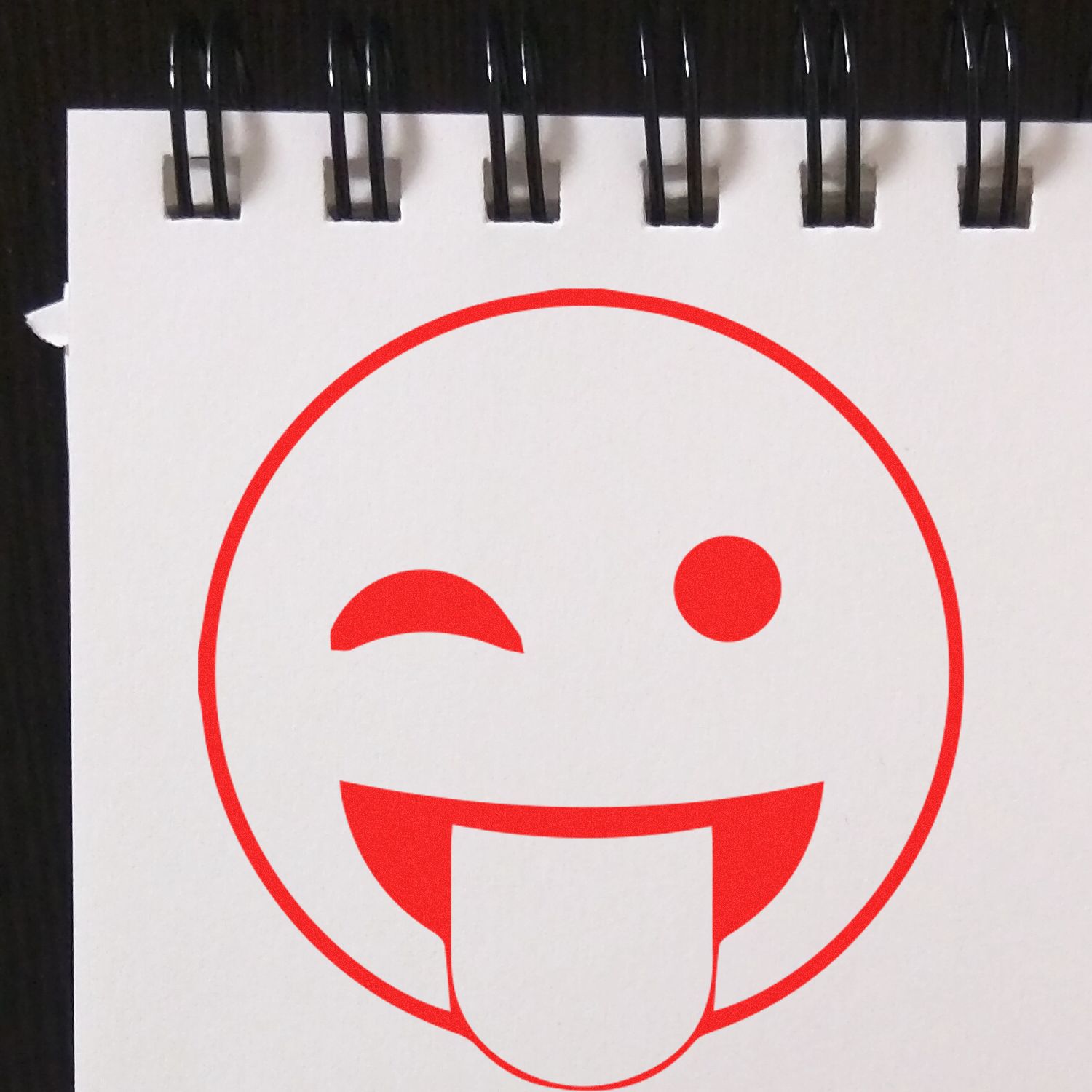 Red winking tongue-out emoji face printed on a spiral-bound sketchbook page, playful graphic design icon.