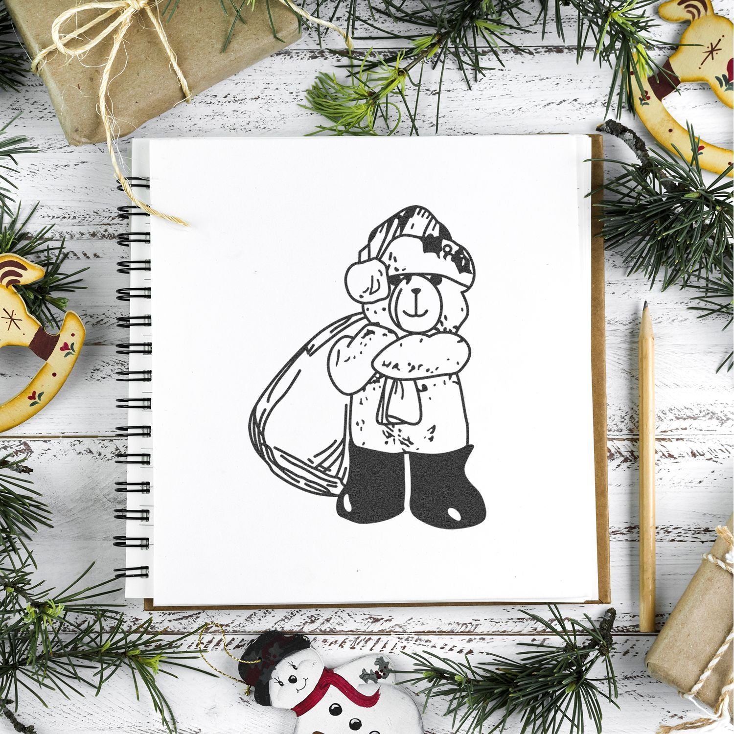 Winter holiday sketchbook with cute bear illustration, surrounded by pine branches, wrapped gifts, and festive ornaments on a rustic white wooden background.