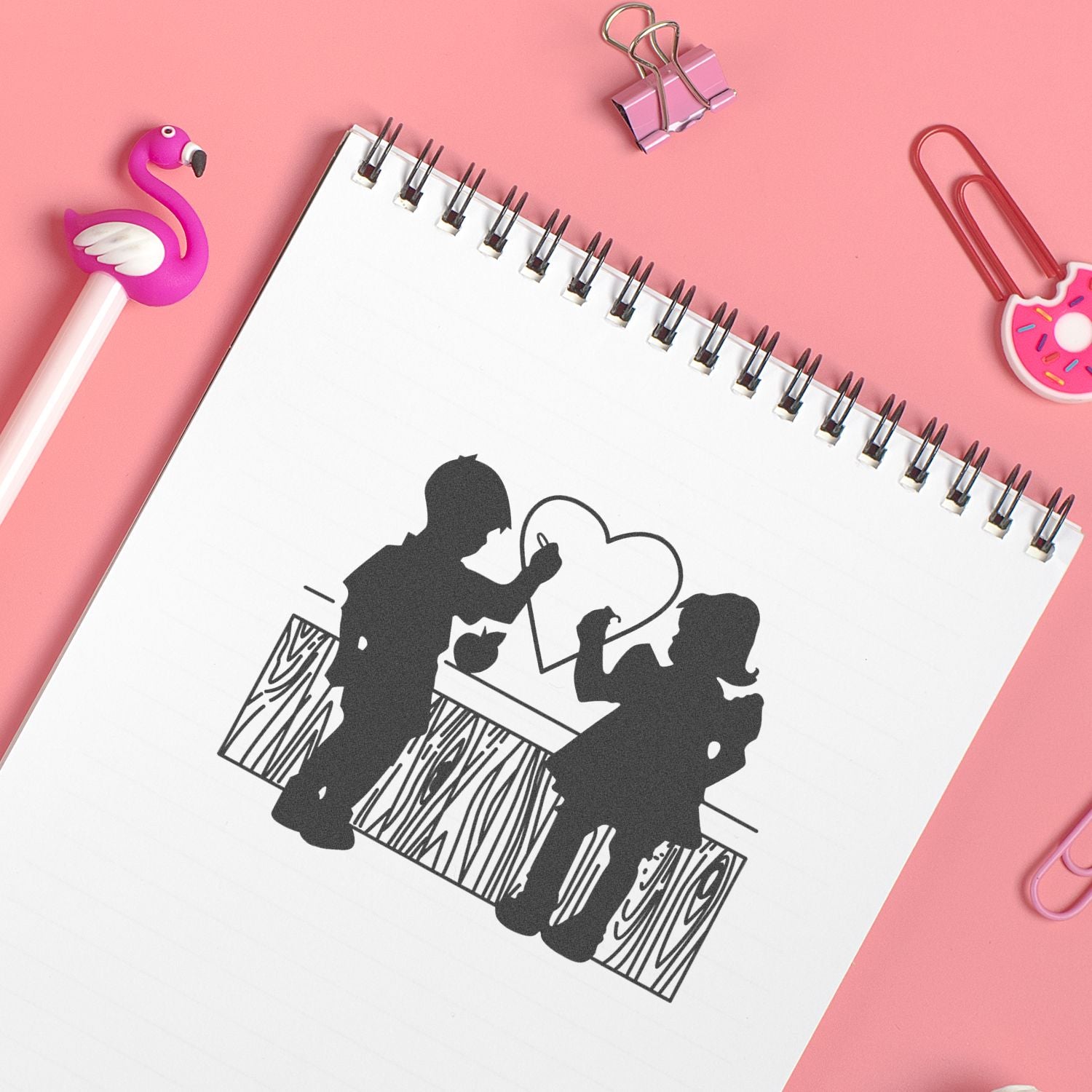 Cute stationery flat lay with spiral notebook featuring children drawing a heart, pink background, flamingo pen, binder clips, and paper clips.