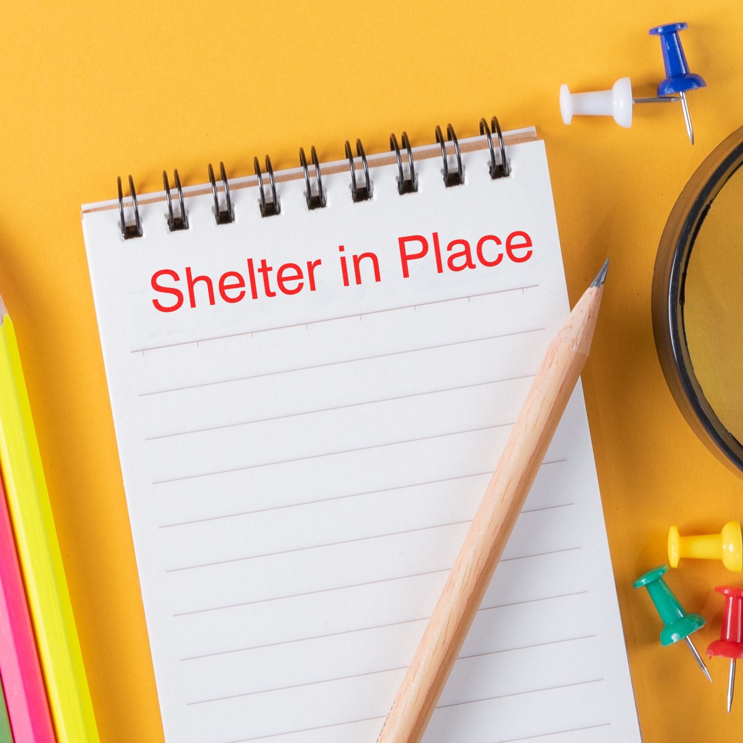 Notepad with Shelter in Place text, pencil, and colorful push pins on a yellow background.