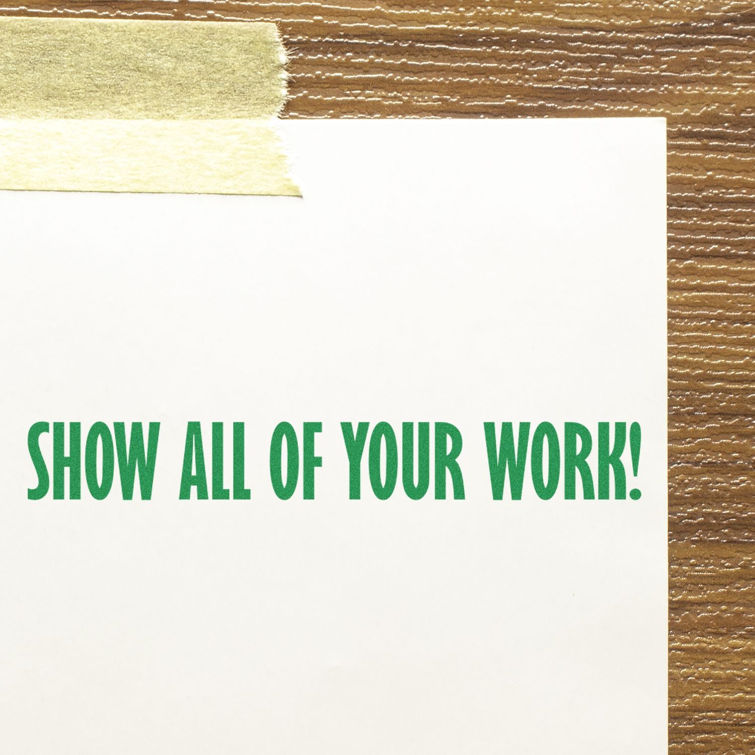 White paper sign with green text reading Show All of Your Work! taped to a brown textured background.
