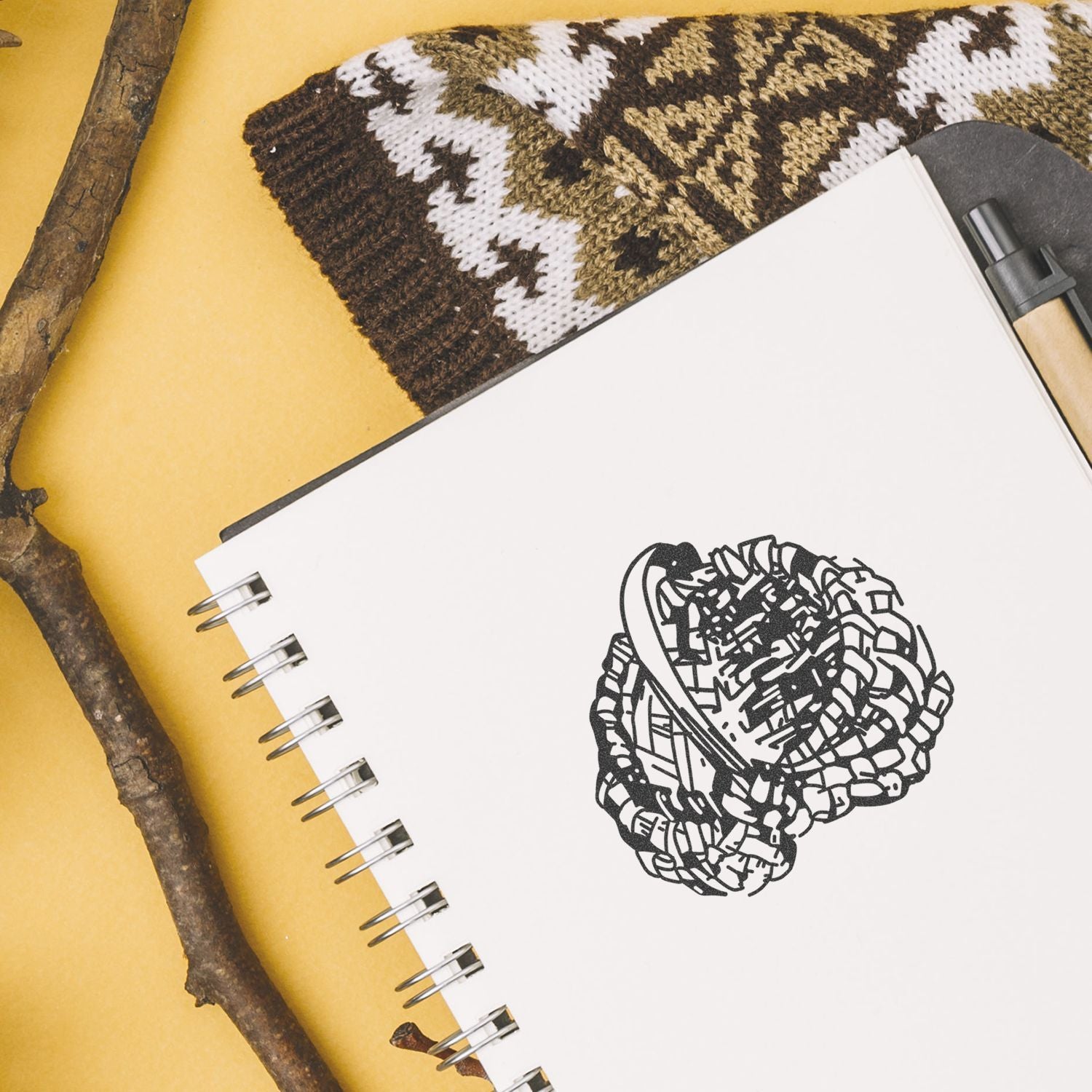 Spiral notebook with abstract black logo, pen, knitted fabric, and rustic branch on yellow background flat lay.
