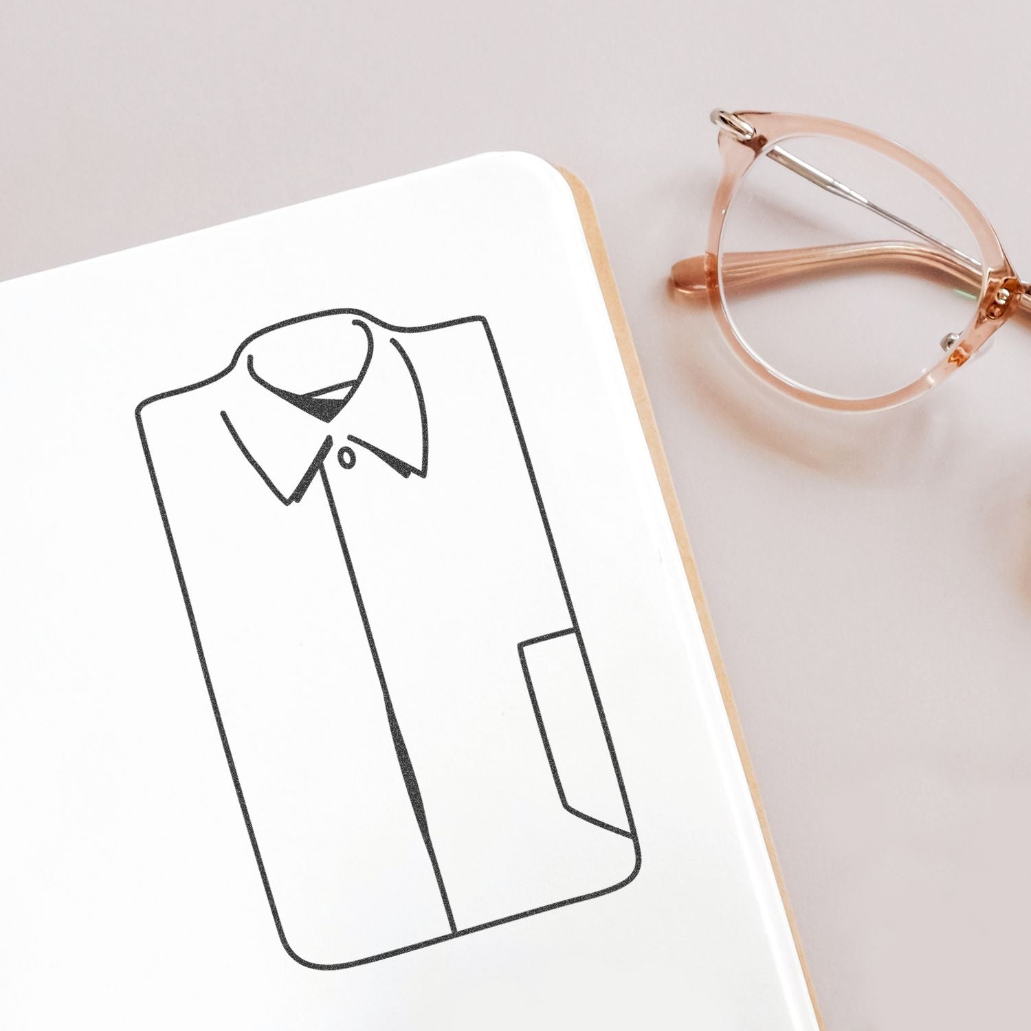 Minimal fashion sketch of a button-up shirt on a notebook beside rose gold eyeglasses.
