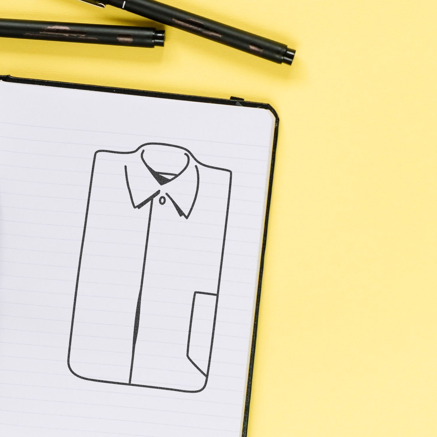 Notebook with a hand-drawn shirt design sketch and black pens on a yellow background, fashion illustration concept.