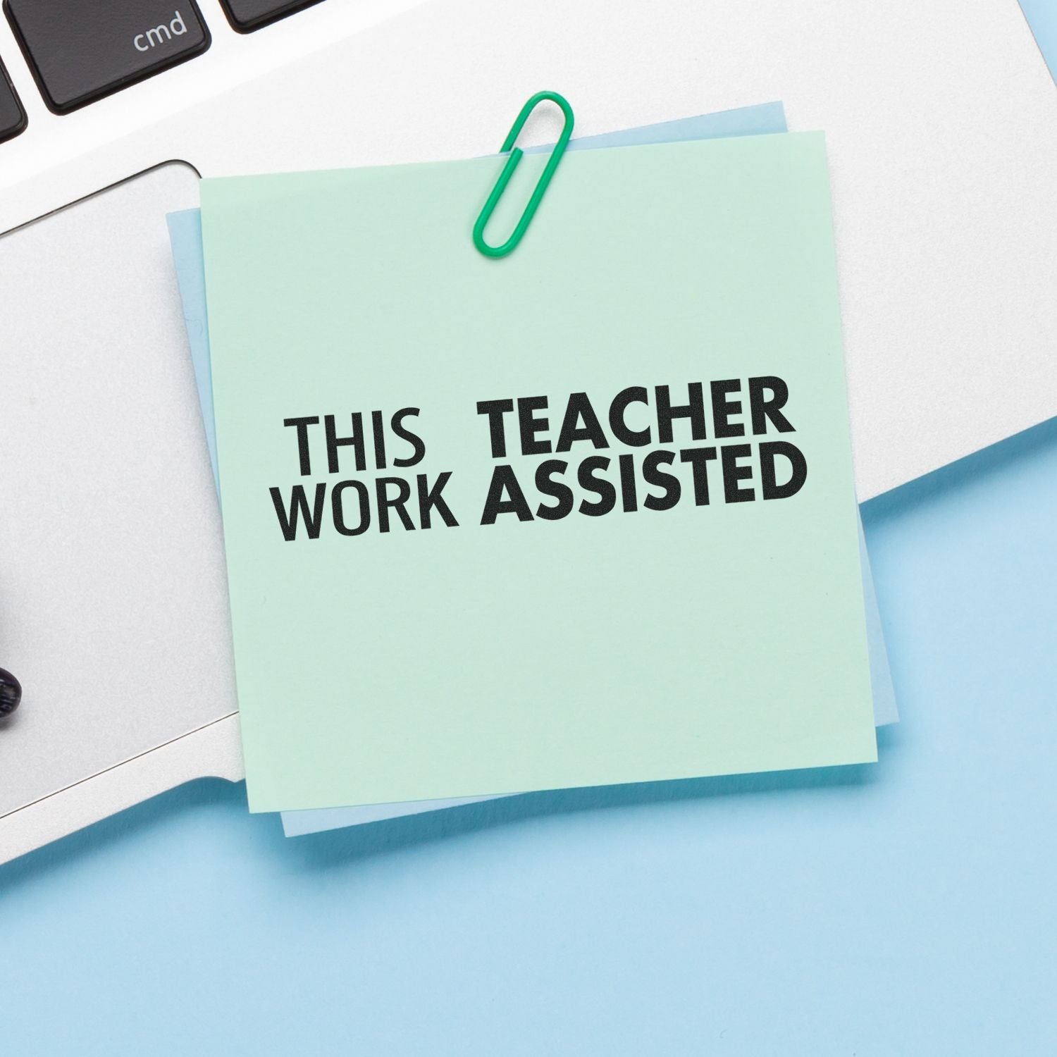 Desk sticky note reading This Teacher Work Assisted with paperclip, highlighting teacher support and education assistance.