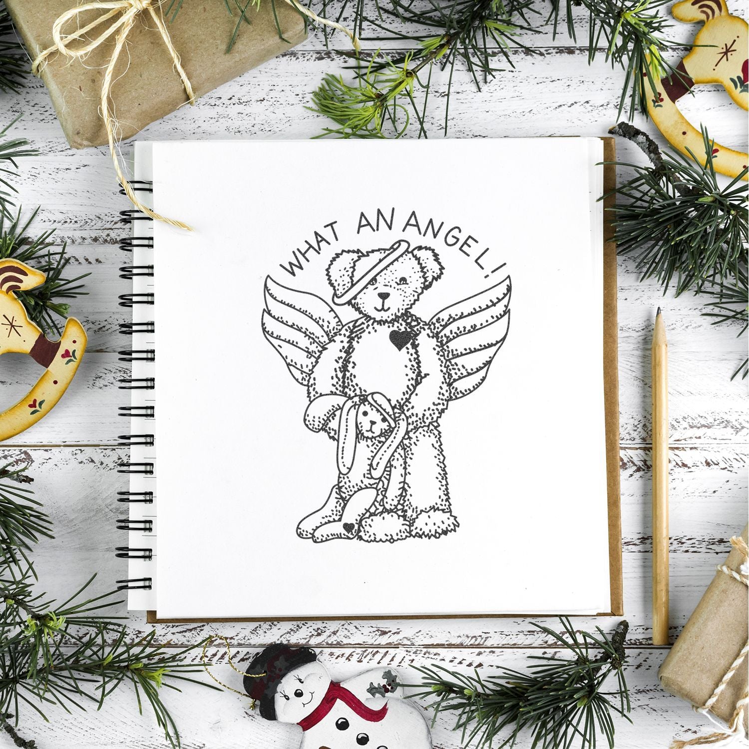 Christmas sketchbook with angel teddy bear illustration, surrounded by pine branches, wrapped gifts, and festive decorations on a rustic white wooden background.
