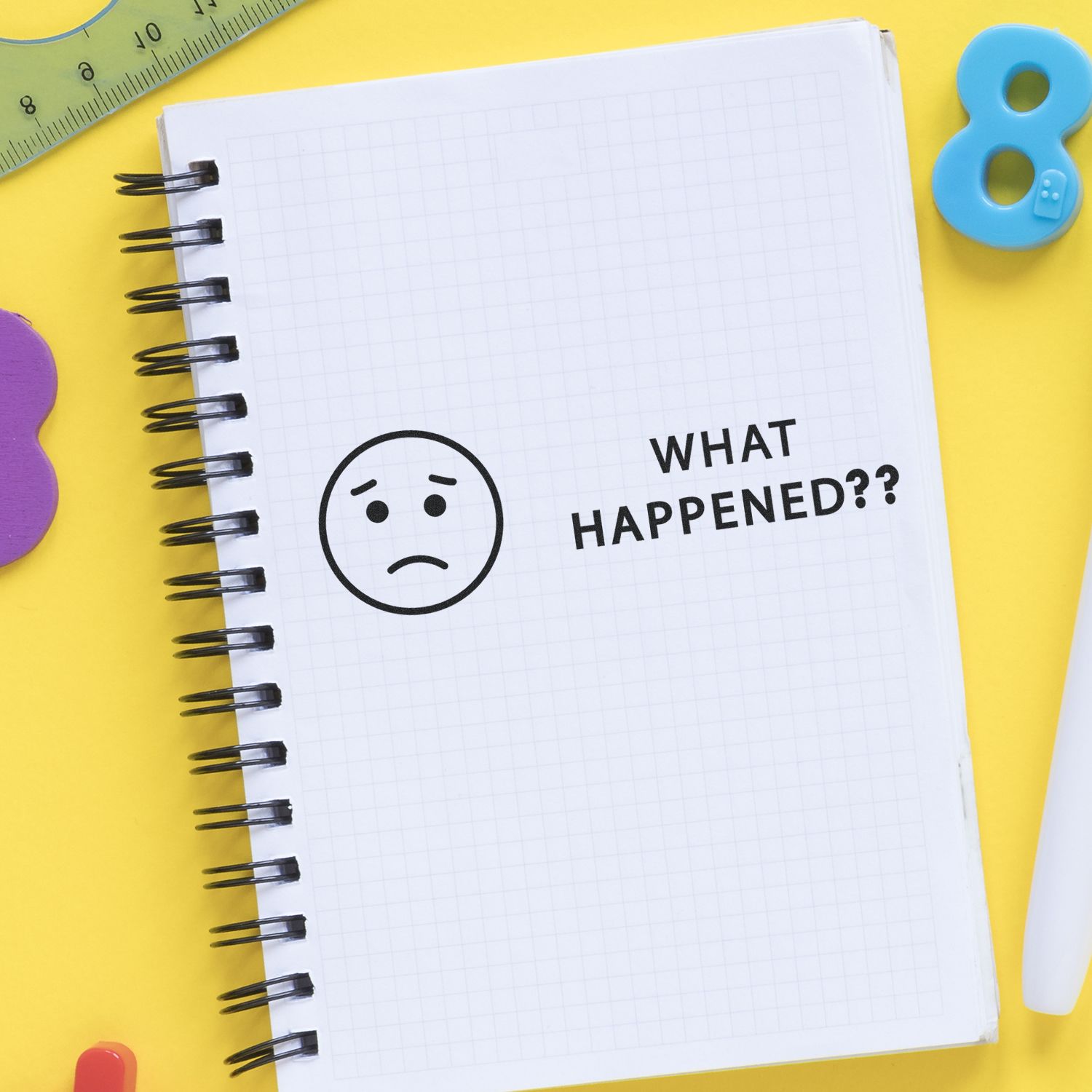 Sad face doodle and What Happened?? text in a notebook on a yellow background with school supplies, creative back to school concept.