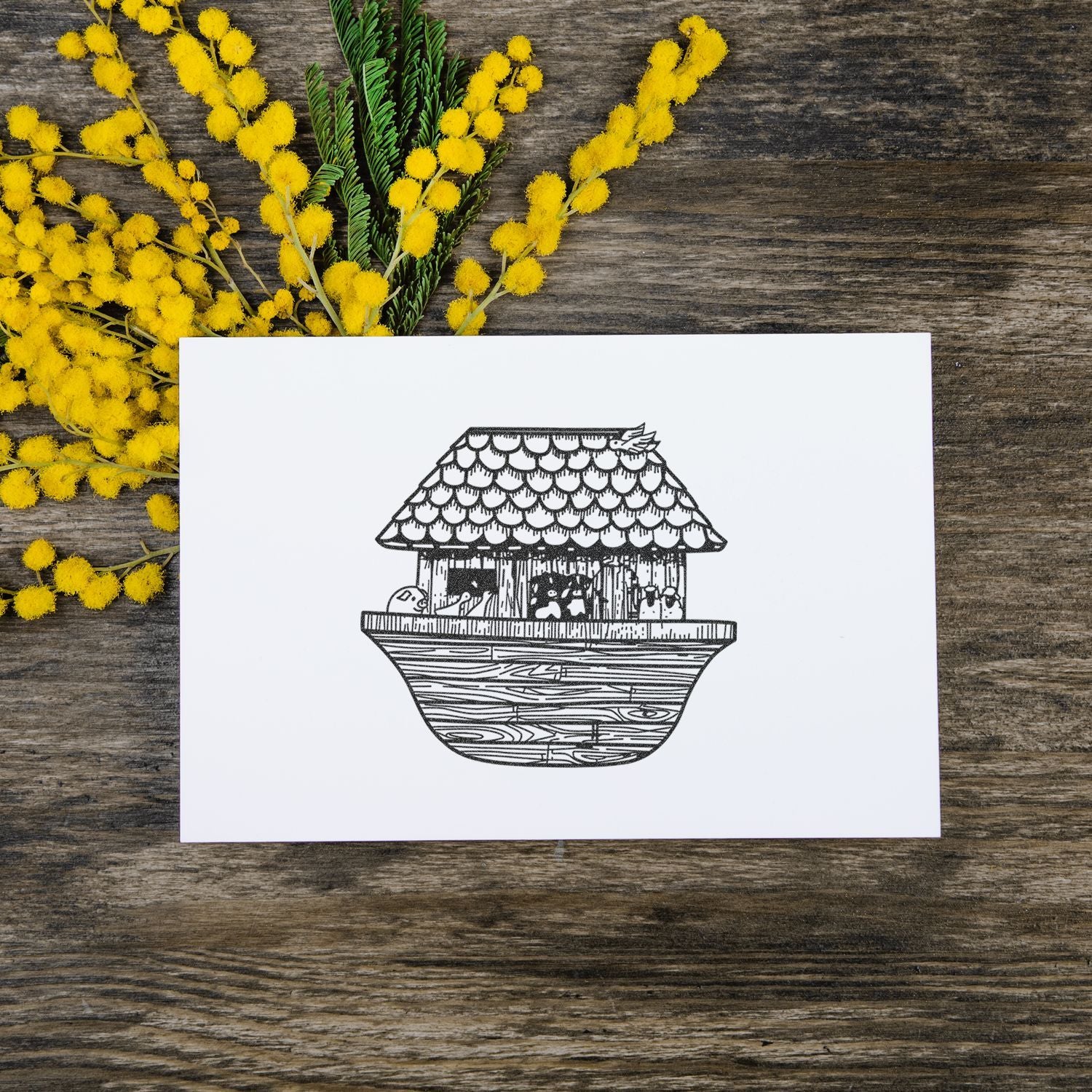 White card with a black line drawing of a small house on water, surrounded by yellow mimosa flowers on a rustic wooden background.